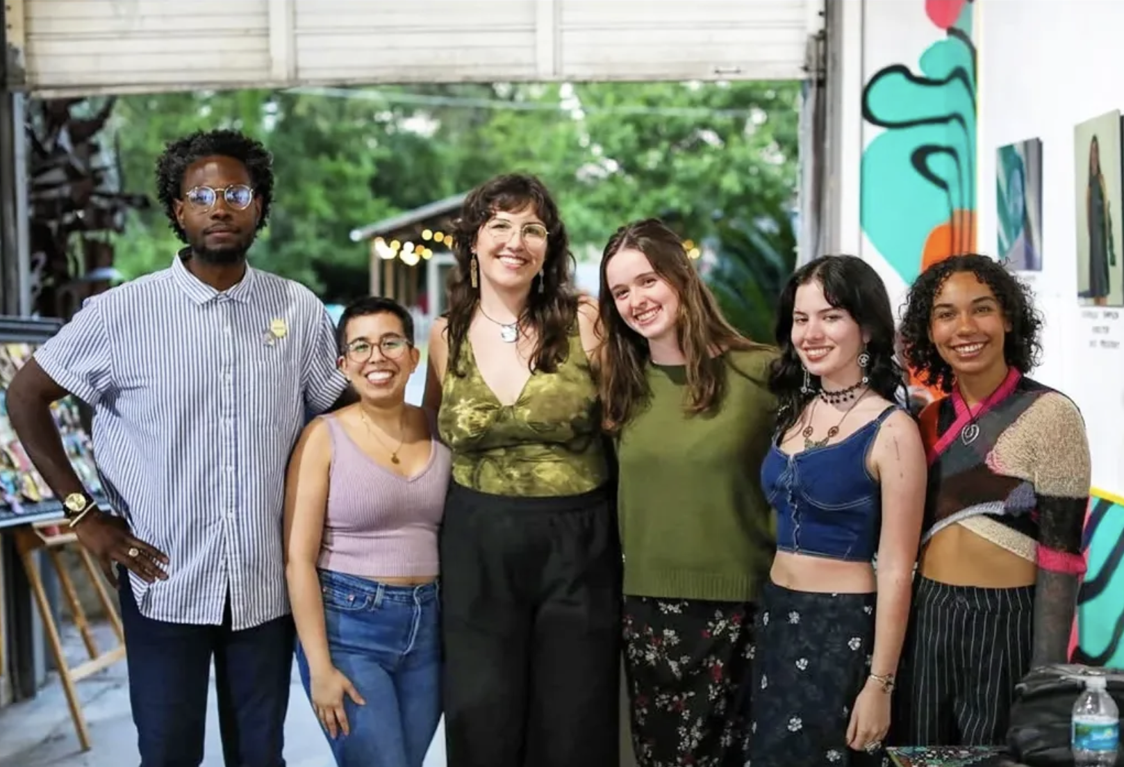 A group of seven diverse women and one man smiling together at an outdoor art exhibit or festival, with artwork and trees visible in the background.