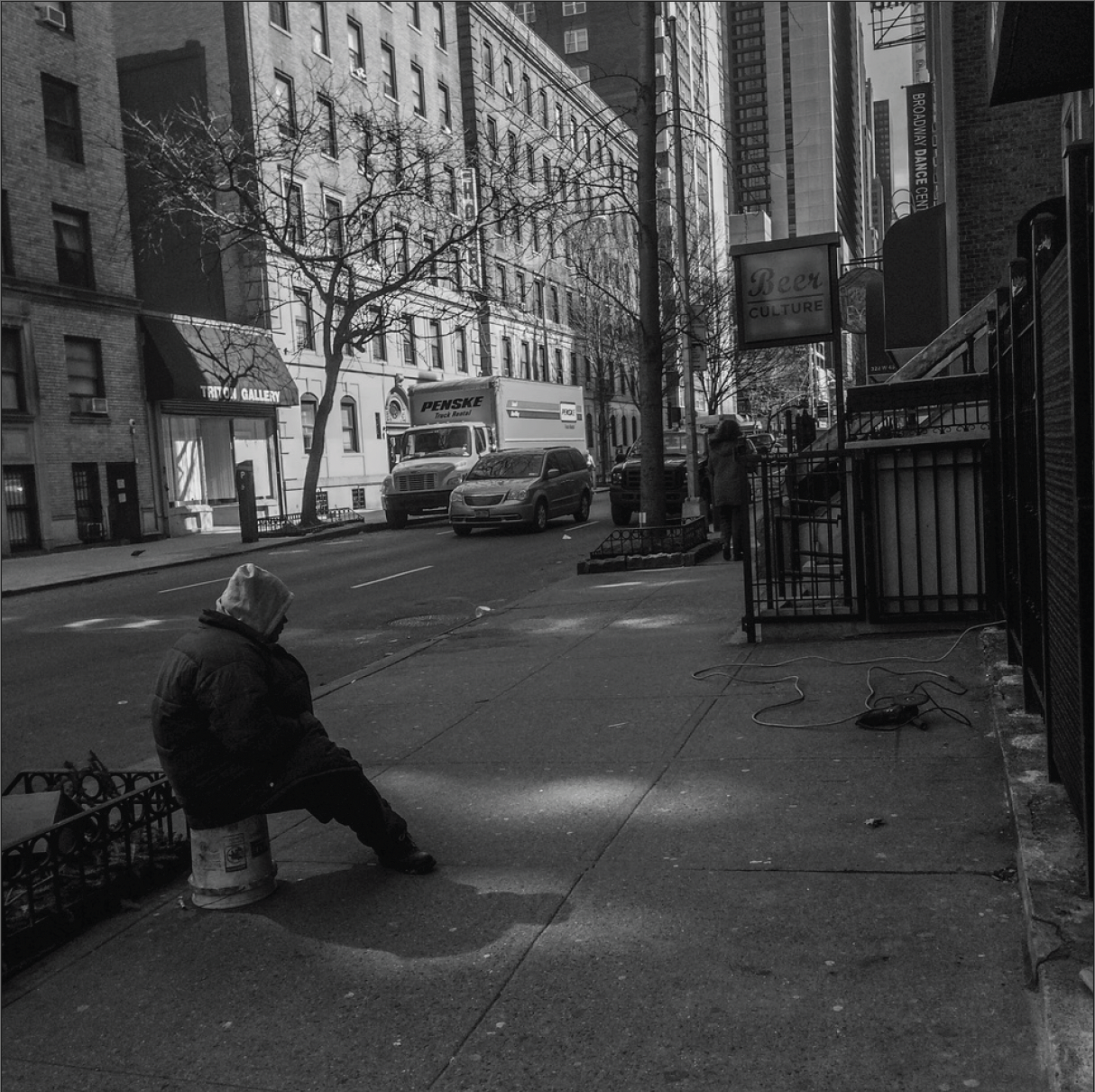 Black and White image of person sits on bucket on city sidewalk
