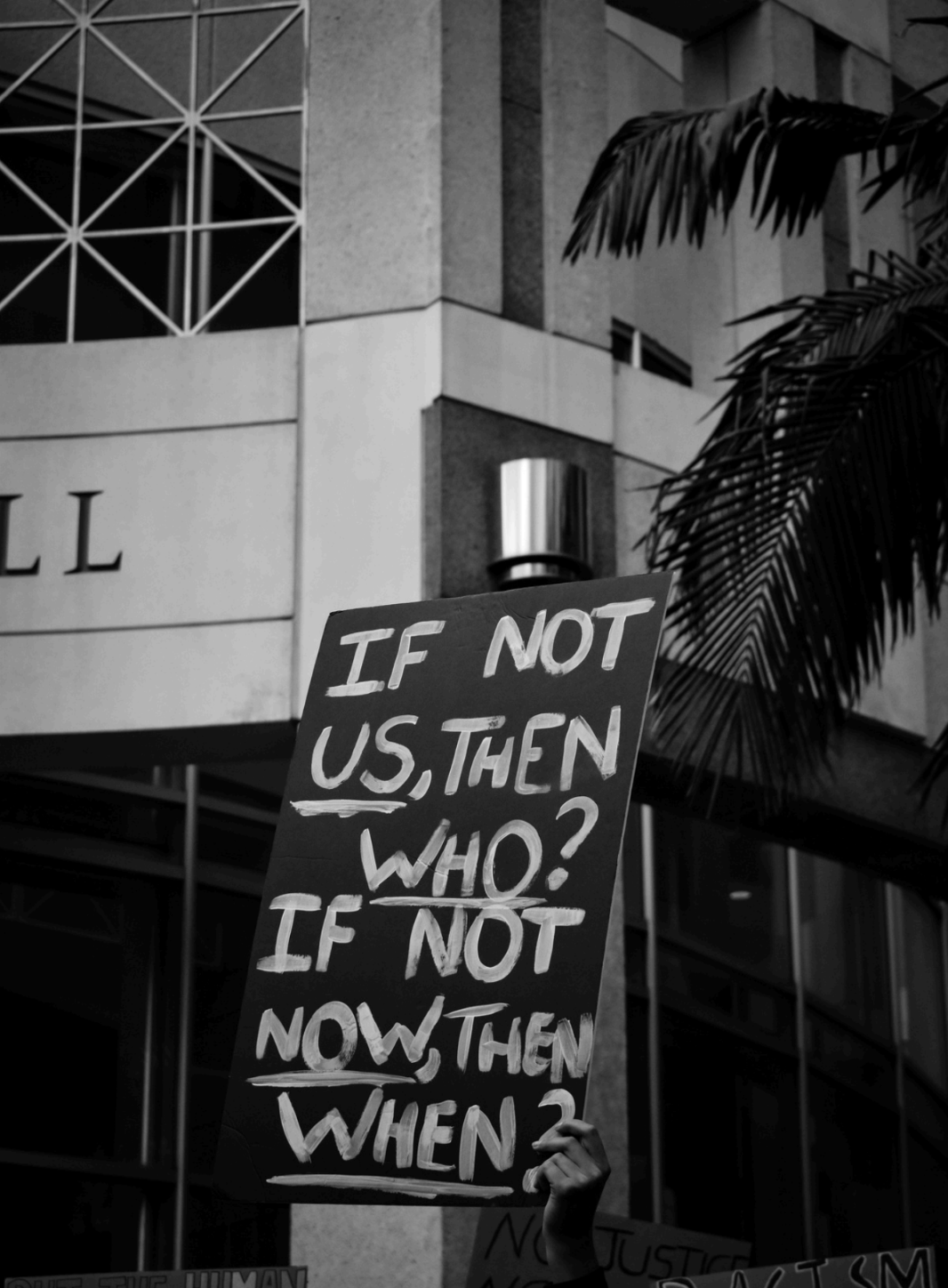 Black and White image of hand holding sign saying, "IF NOT US, THEN WHO? IF NOT NOW, THEN WHEN?"