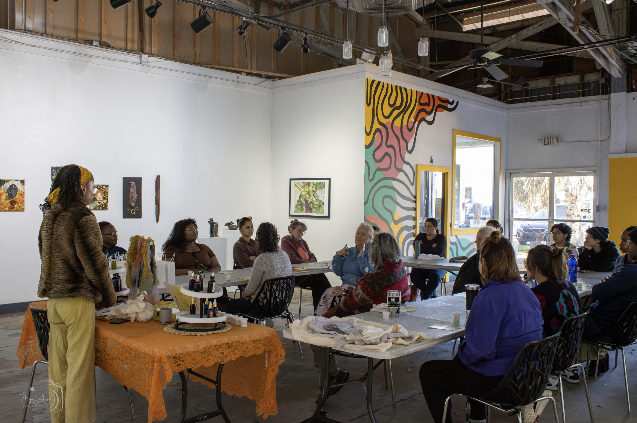 People gathered in a brightly lit art studio with colorful wall murals, seated around tables and listening to a woman standing, who is giving a demonstration.