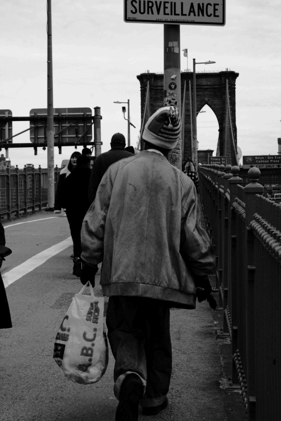 Black and White image of person walking with a white tote back in hand on a bridge as other pedestrians walk in the background