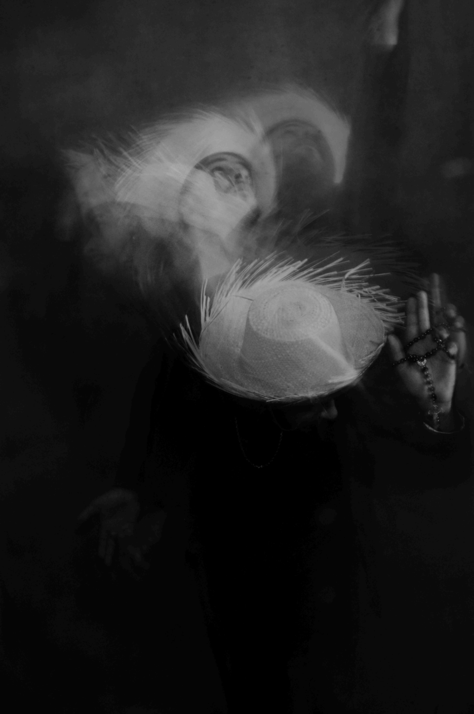 Black and White blurred image of person in straw hat looking downward holding beaded necklace in hand