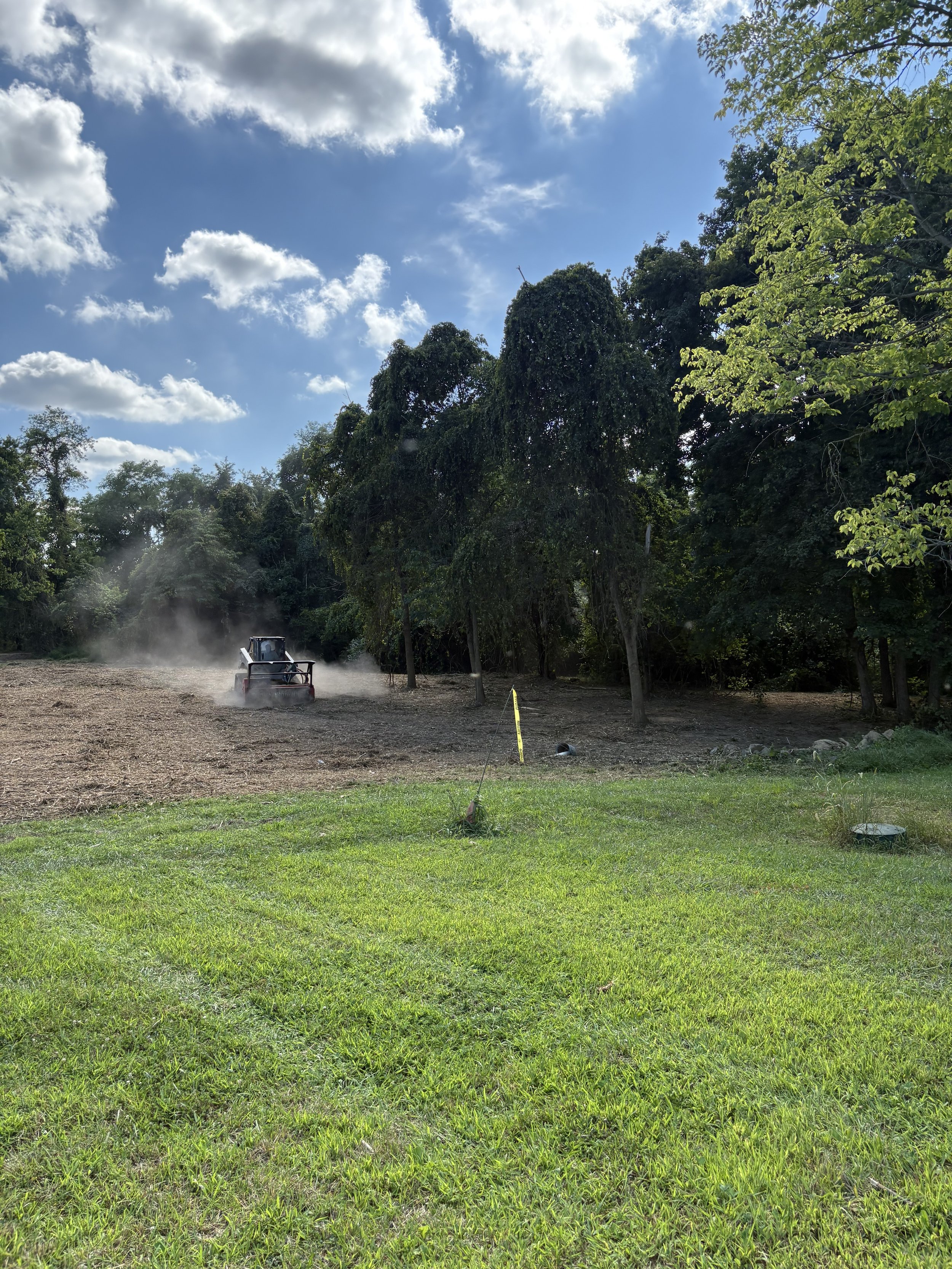 A tractor is tilling or preparing a field near a dense tree line on a sunny day with partly cloudy skies.