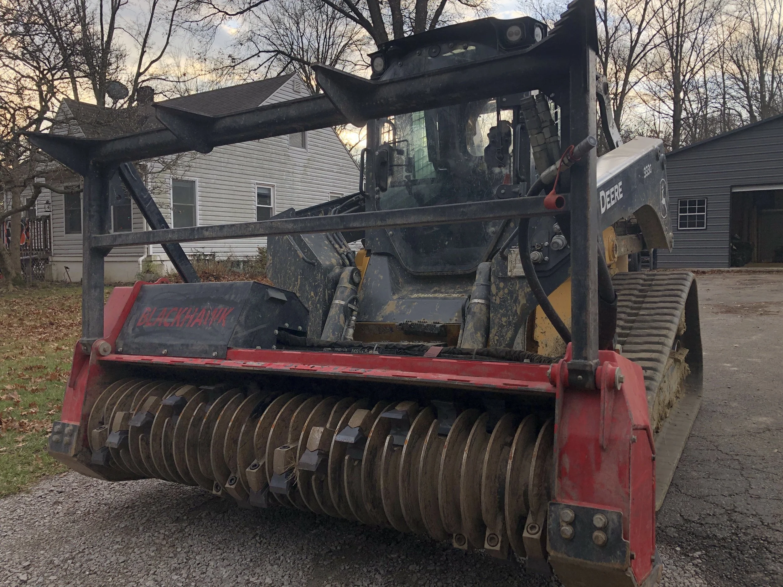 A John Deere construction vehicle with a BlackHawk attachment in a residential area during sunset.