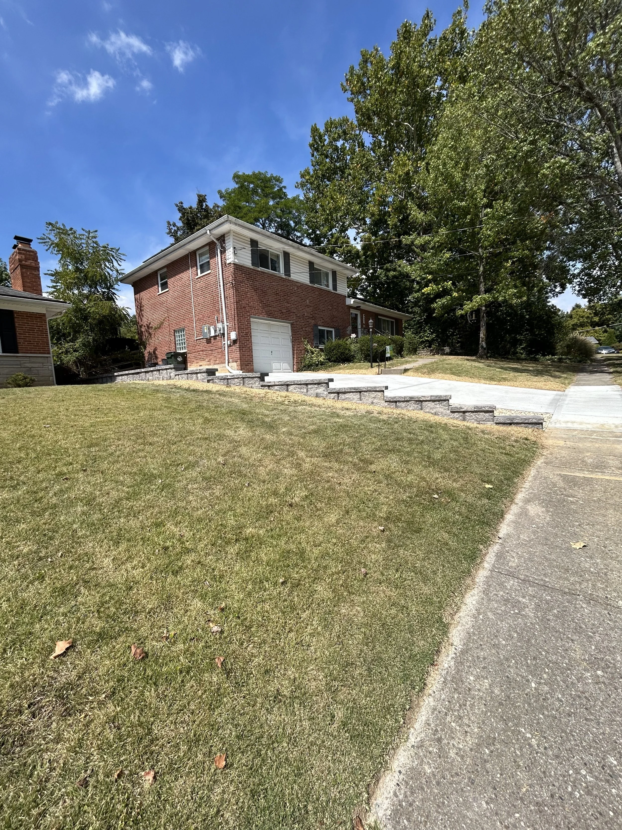 A brick house on a sloped lawn with a concrete driveway and stone retaining wall, surrounded by trees under a blue sky.