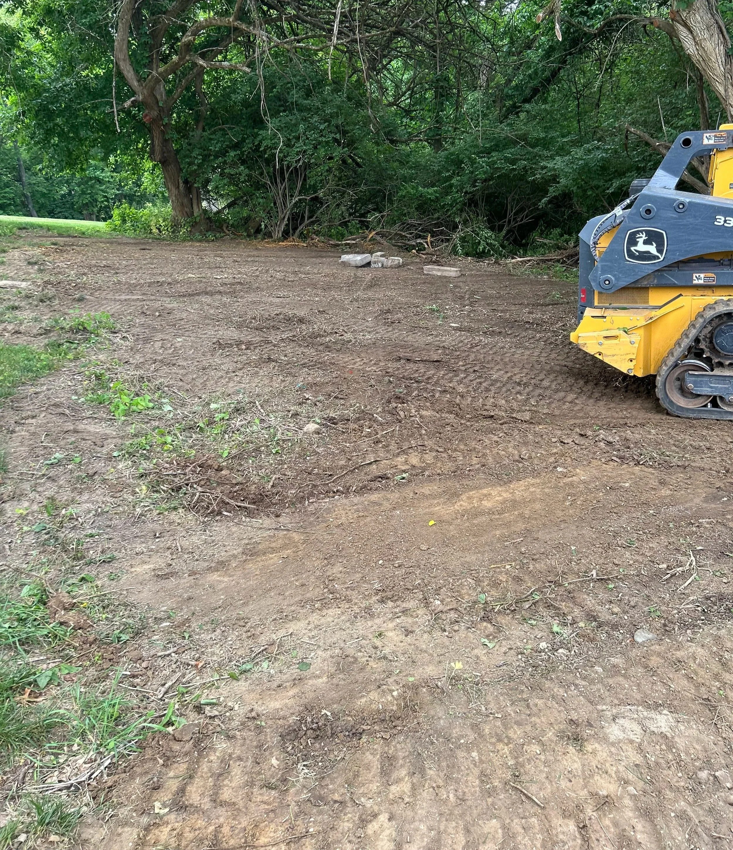 A construction site with cleared dirt ground, some green plants on the edge, and a small yellow tracked construction vehicle on the right side. There are trees and dense greenery in the background.