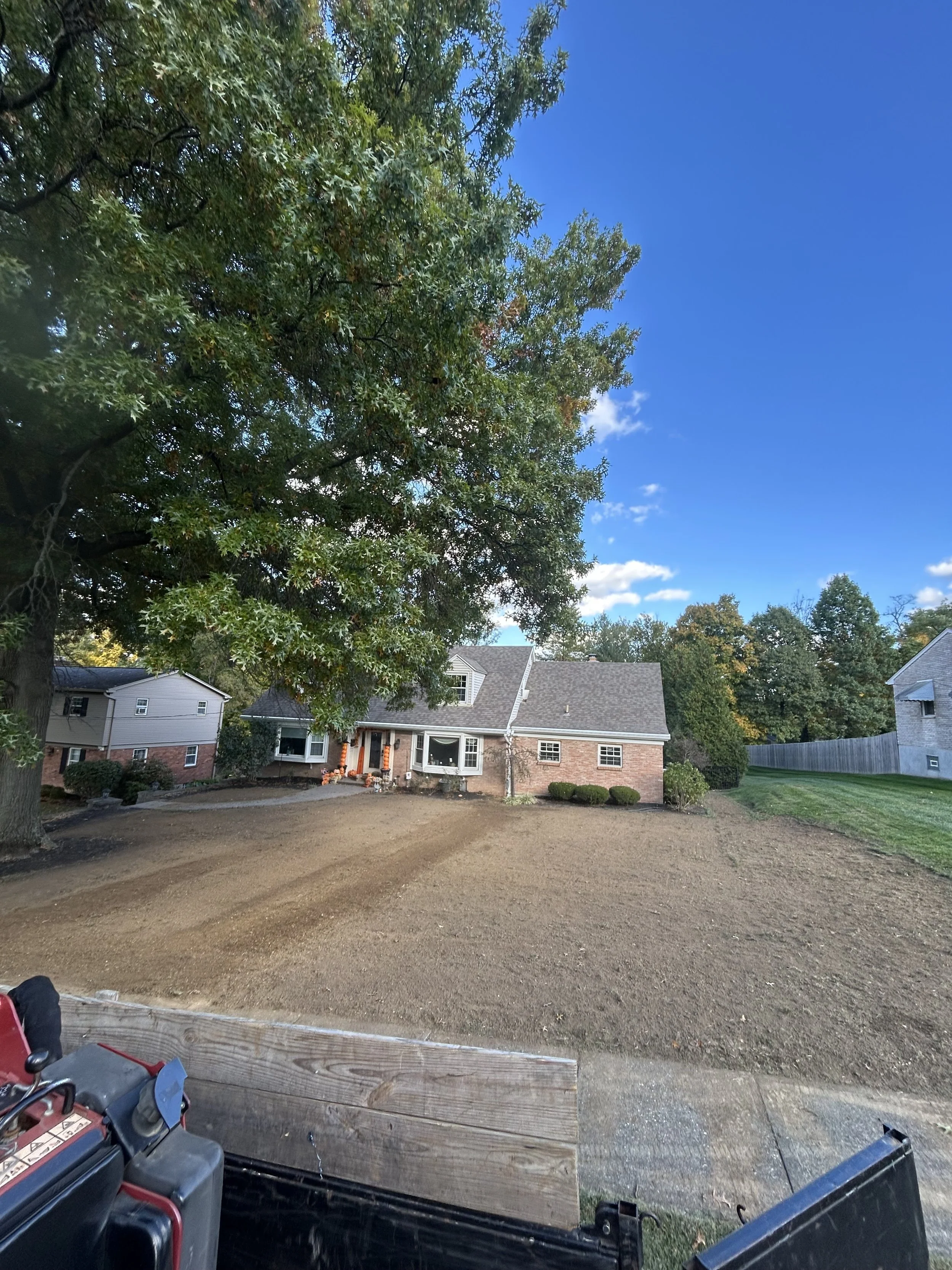 A residential backyard with a large tree on the left, a brick house with a grey roof in the center, and a lawn on the right. The yard appears to be freshly leveled with soil, and a wooden board is visible in the foreground.
