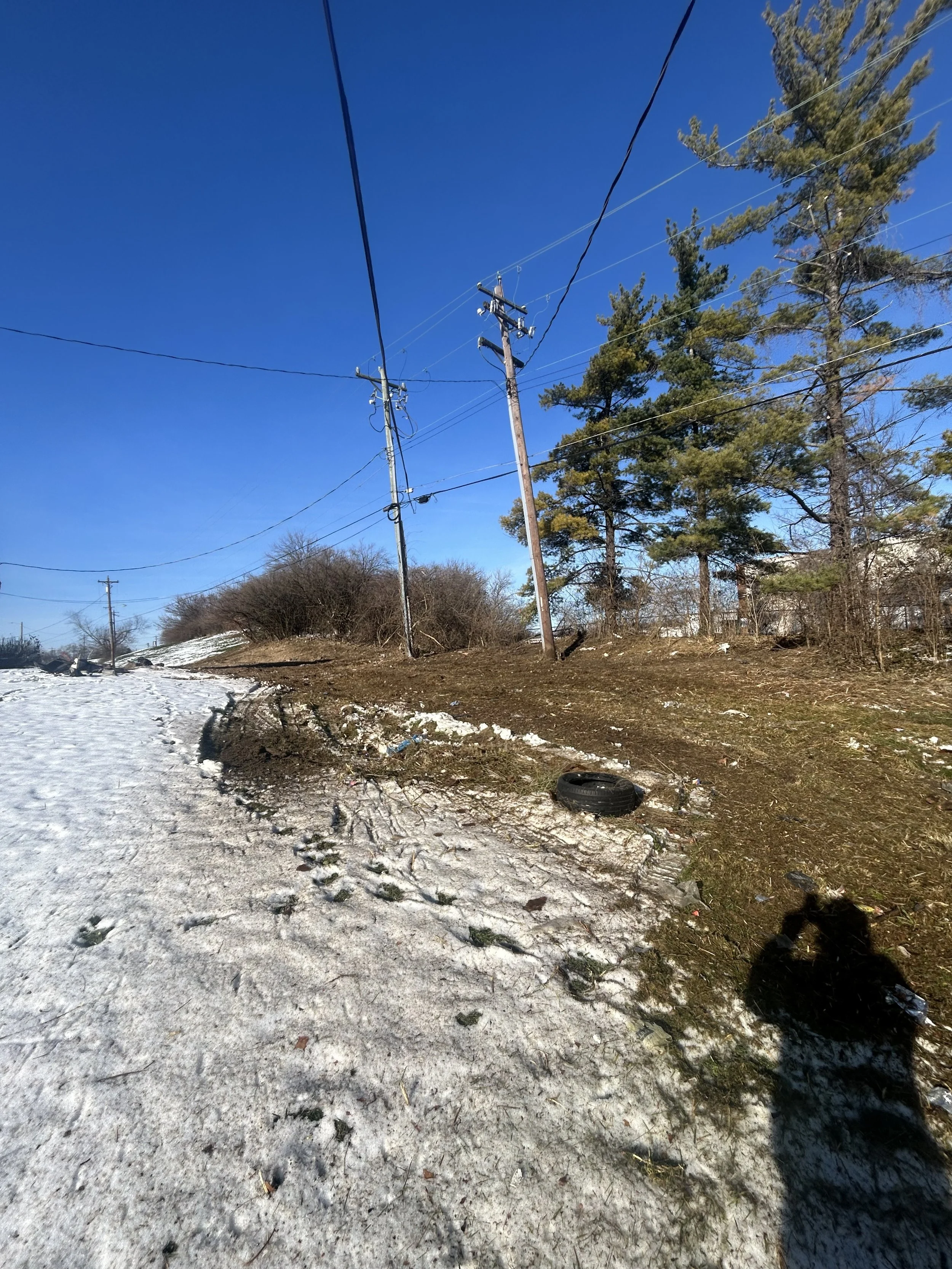 A snowy roadside with a muddy patch, two electric poles with wires, a tire on the ground, and sparse trees under a clear blue sky.