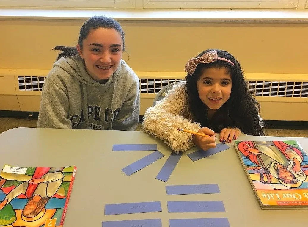 Two young girls sitting at a table, working on a literacy activity with blue strips of paper and books titled 'The Little Girl.'