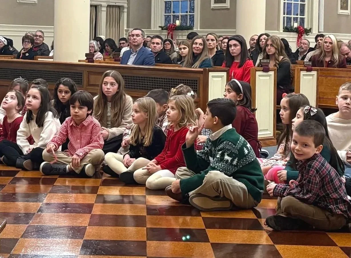 Children sitting cross-legged on a checkered wooden floor in a church setting, with adults seated behind in pews, during what appears to be a festive holiday event.