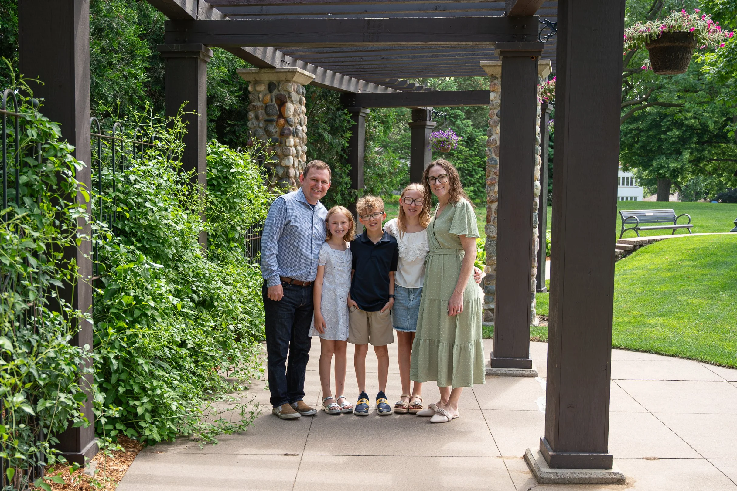 A family of six standing under a wooden pergola in a park, with greenery and a bench in the background, smiling at the camera.