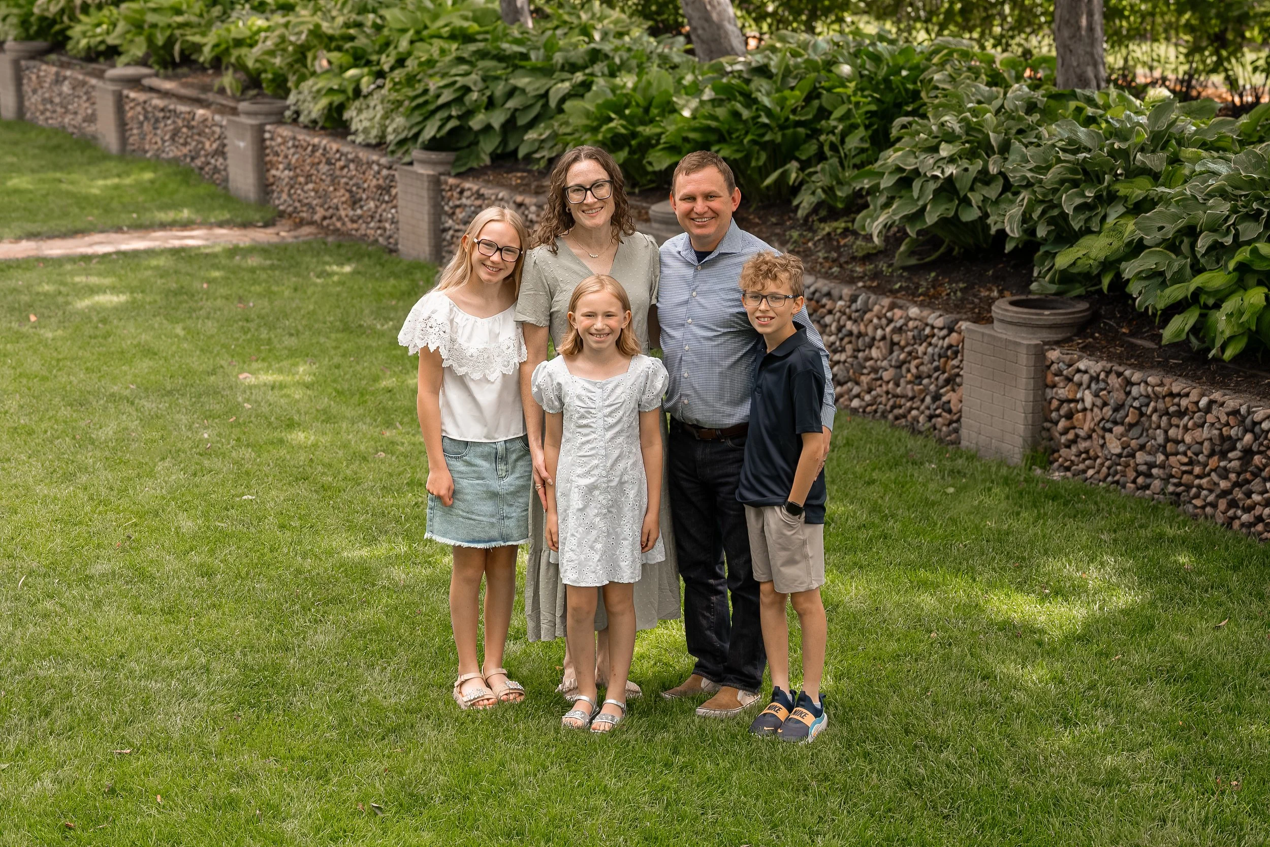 A family of six standing outdoors on a grassy lawn in front of a flower bed with green plants and trees, smiling for a group photo.
