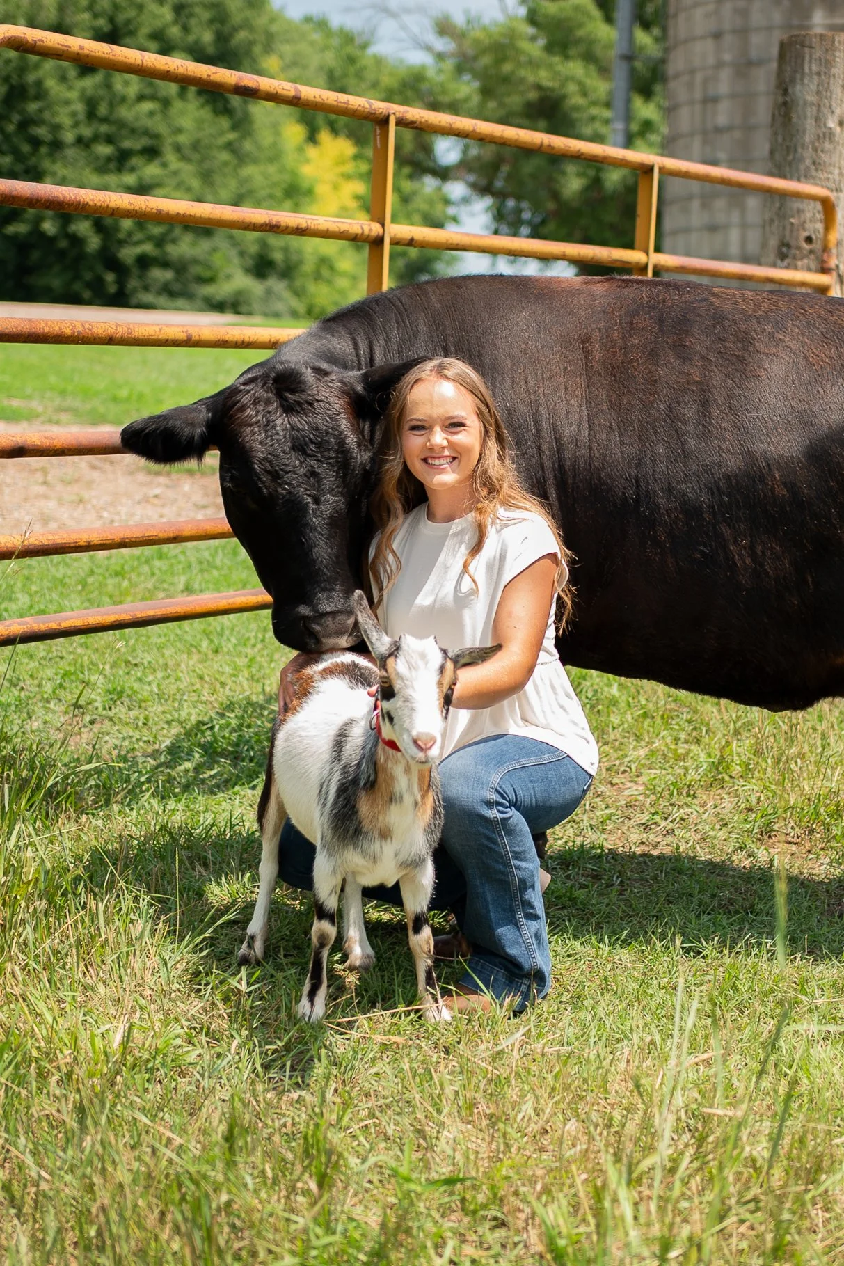 A woman kneeling on grass with a black goat and a small goat, smiling at the camera, with fenced area and trees in the background.