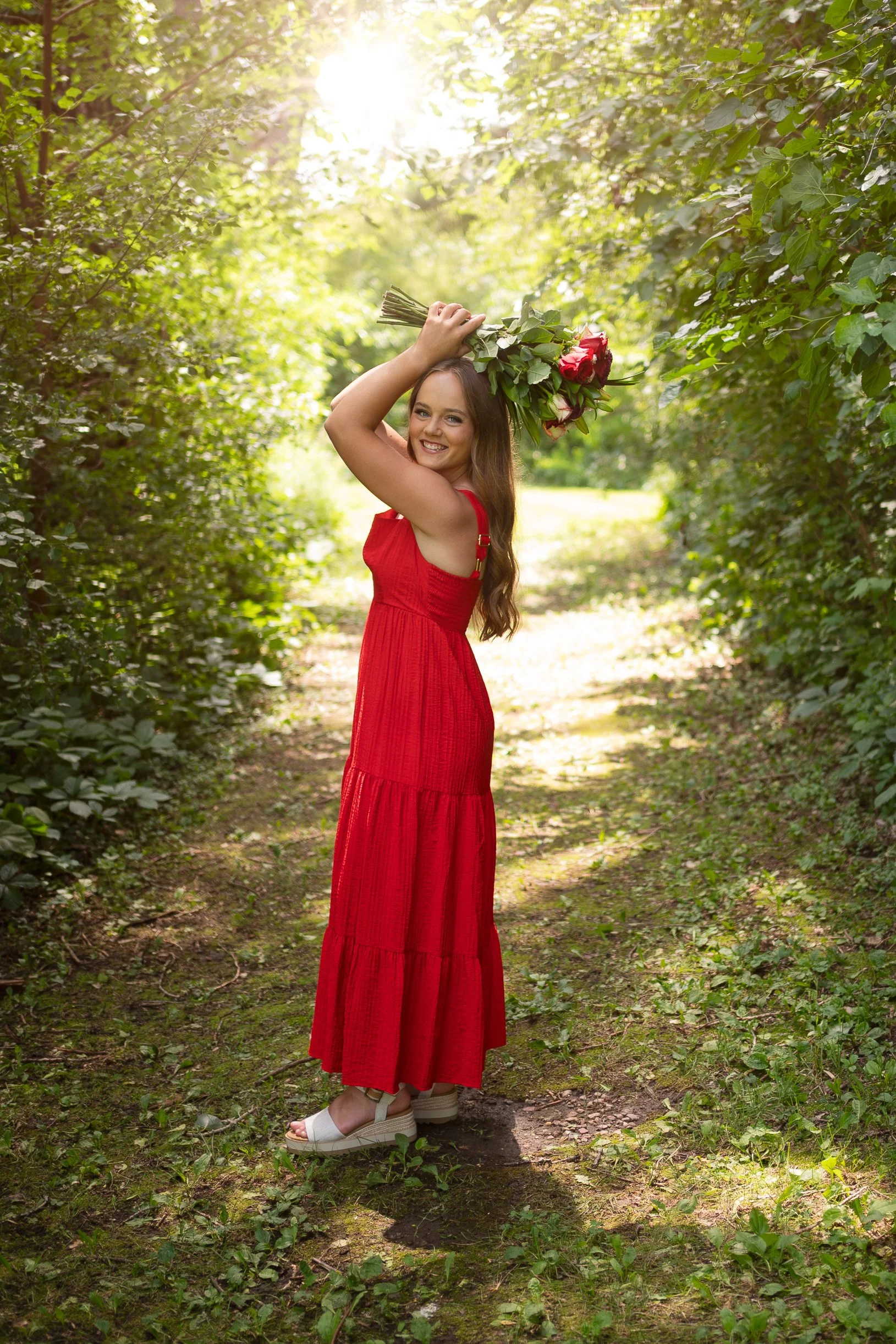 A smiling woman in a red dress holding a bouquet of flowers above her head in a forested area with sunlight filtering through the trees.