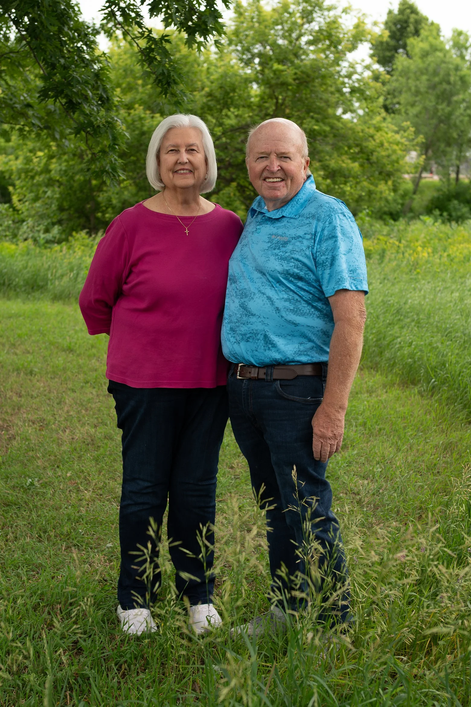 An elderly woman with gray hair and a necklace with a cross, wearing a magenta top, standing next to an elderly man with a bald head, wearing a bright blue patterned shirt, outdoors in a green park with trees and grass.