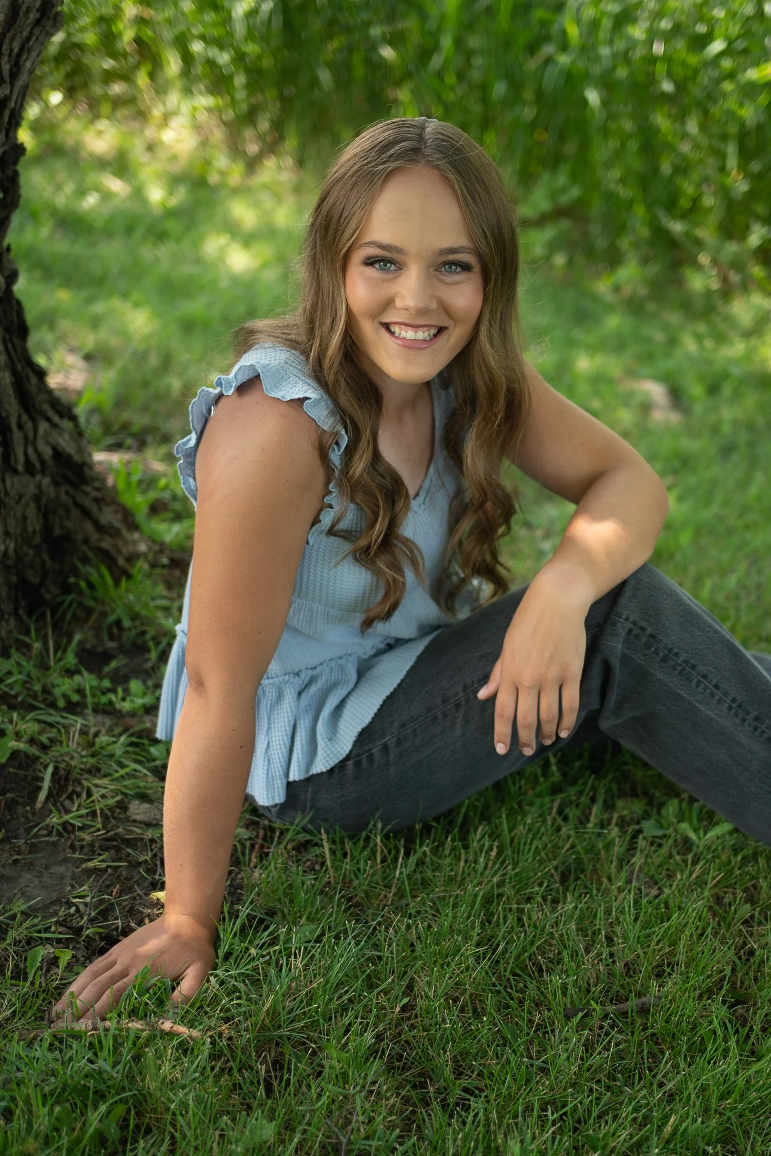 A young woman with long wavy brown hair and blue eyes sitting outdoors on grass near a tree, smiling at the camera.