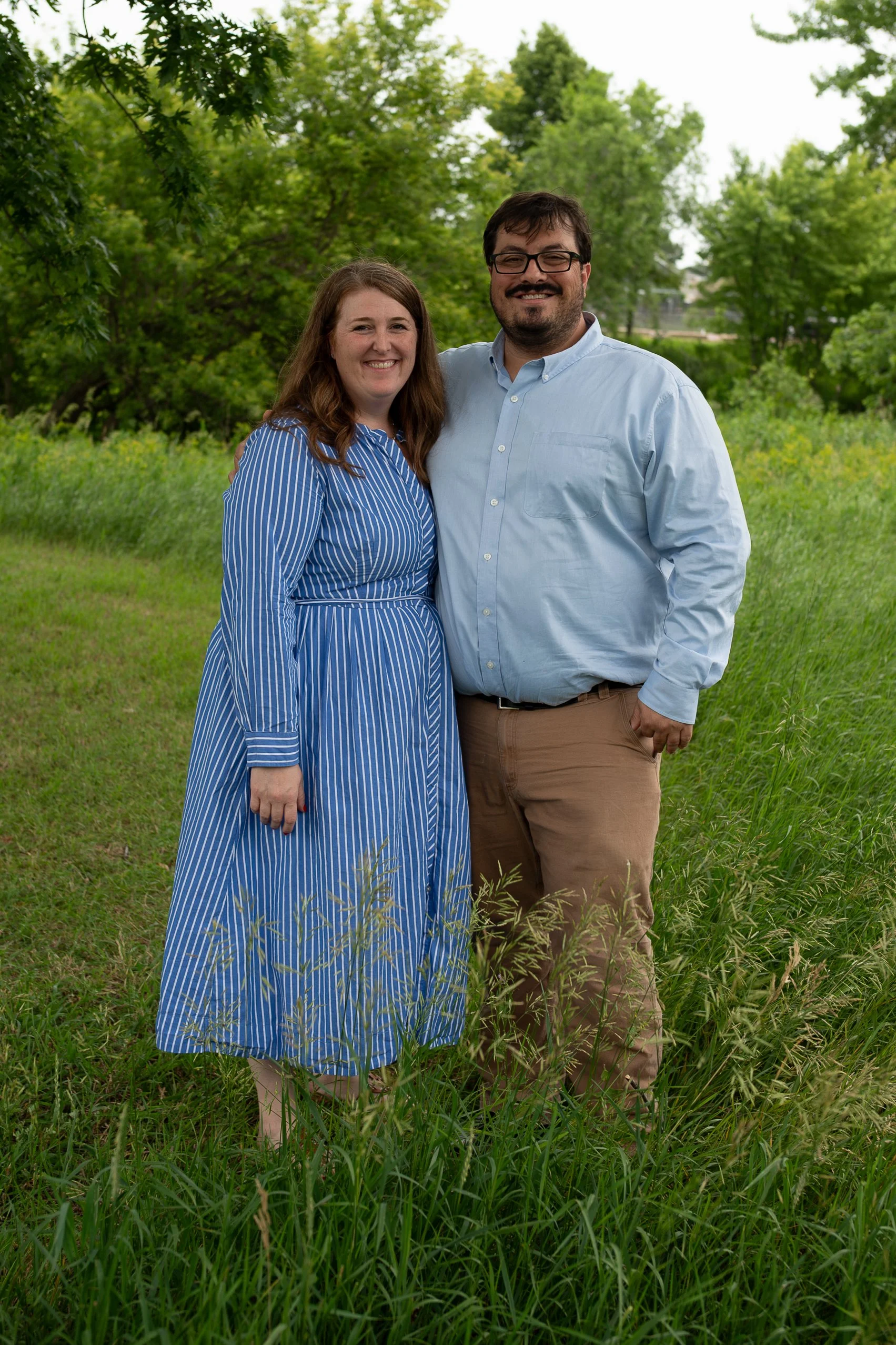 A smiling couple standing in a grassy field with green trees in the background. The woman is wearing a blue and white striped dress, and the man is wearing a light blue button-up shirt and khaki pants.