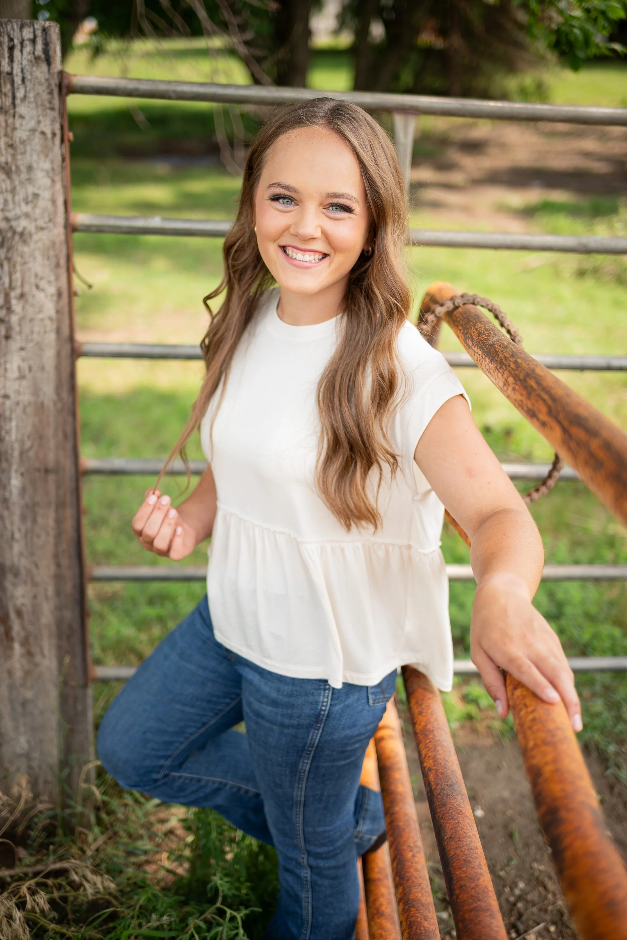 A smiling woman with long brown hair, light skin, wearing a white sleeveless top and blue jeans, standing outdoors near a rusty metal railing and wooden post, with green grass and trees in the background.