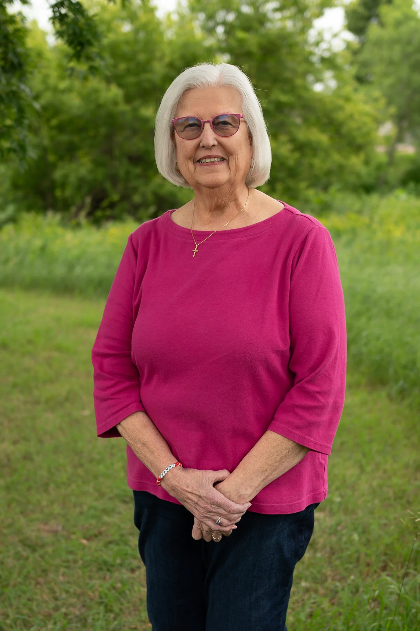 An elderly woman with white hair and glasses, wearing a pink top and dark jeans, standing outdoors with greenery in the background.