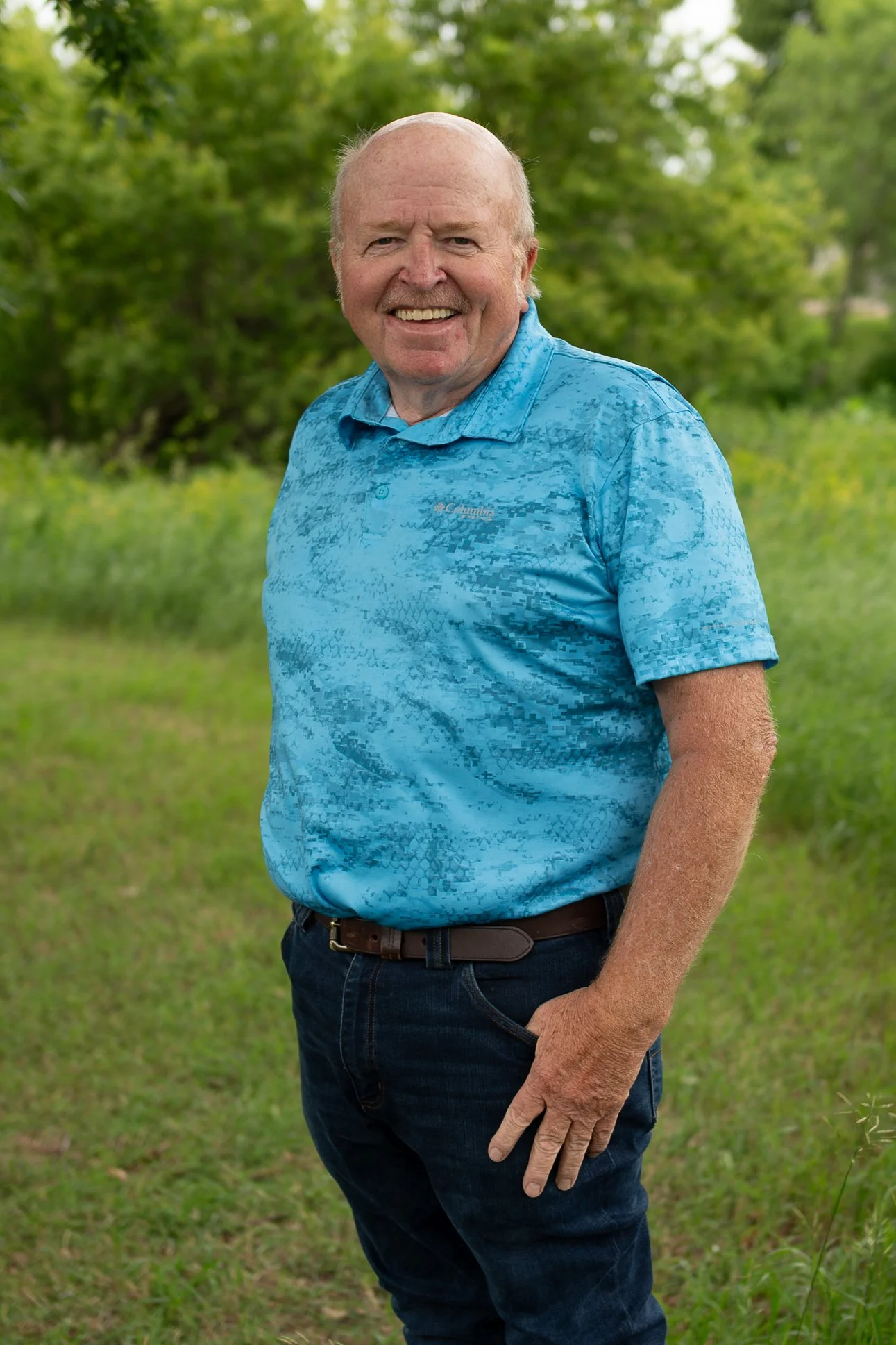 A senior man with a bald head and a big smile is standing outdoors on a grassy field, wearing a blue patterned short-sleeve shirt and dark pants, with green trees and foliage in the background.