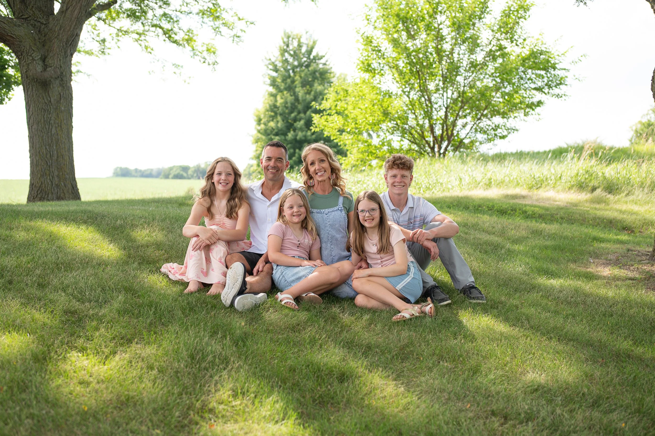 A family of six sitting on the grass under a tree in a park on a sunny day, smiling at the camera.