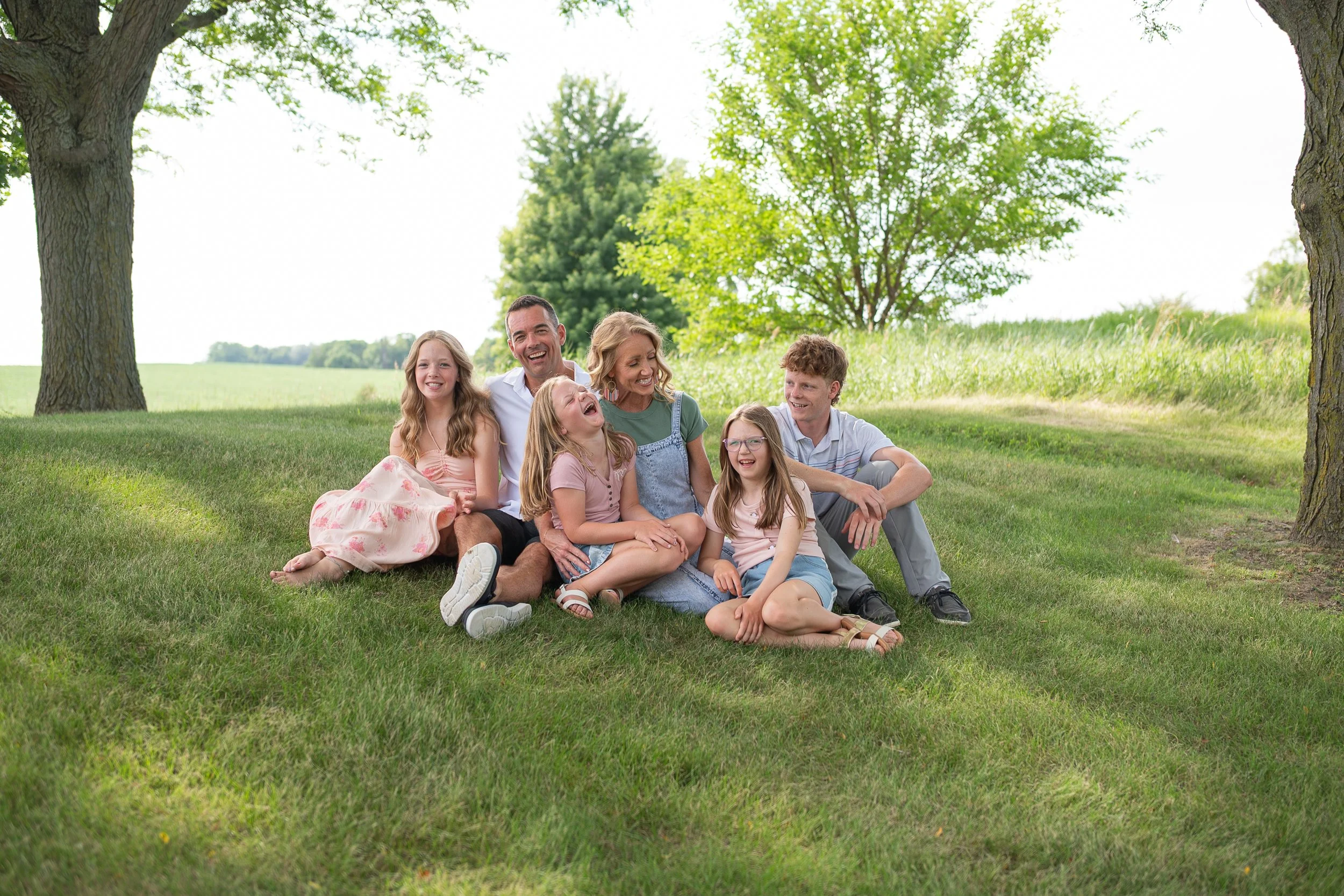 Family of seven sitting on the grass under trees, laughing and enjoying a sunny day outdoors.