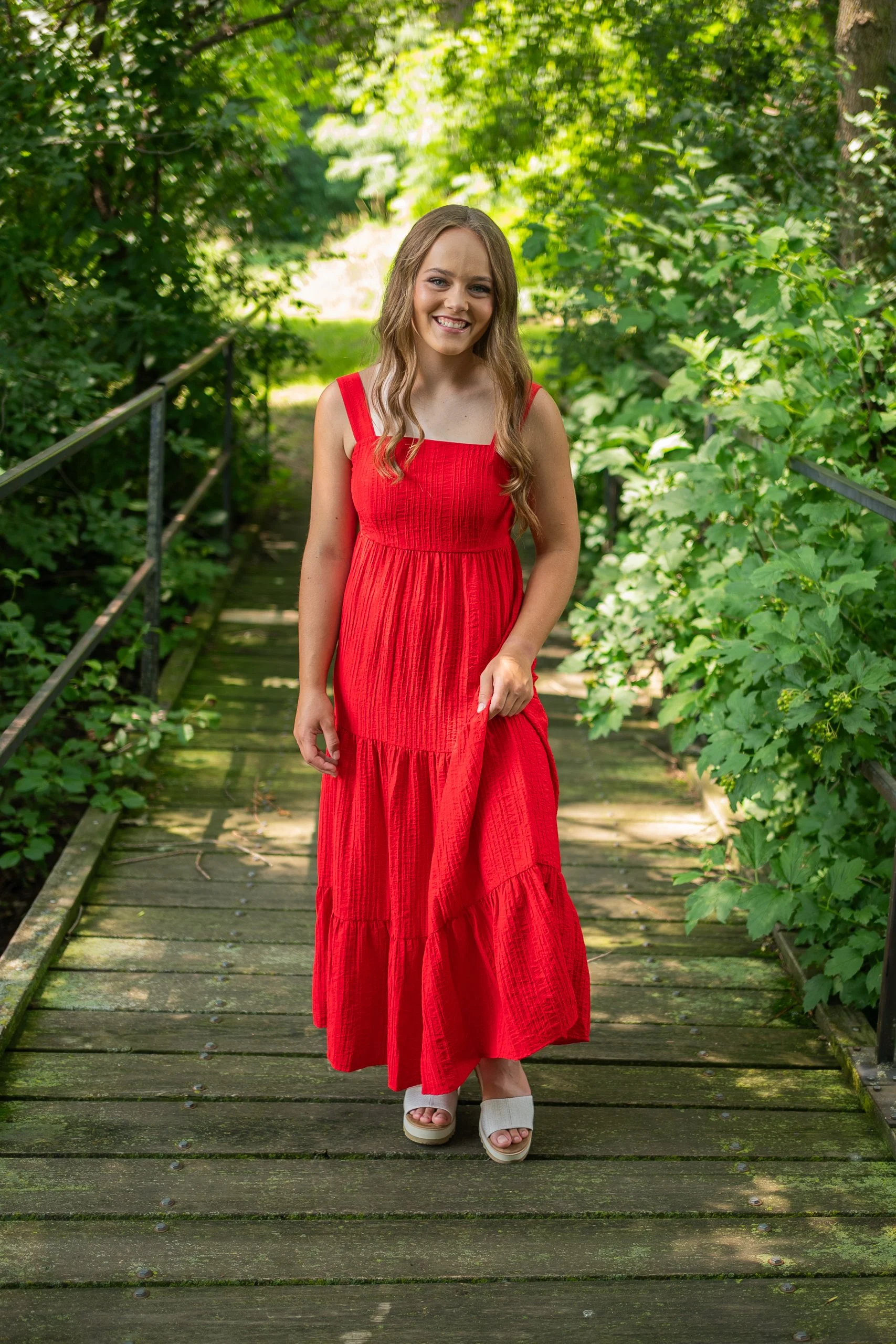 A young woman with long light brown hair, smiling, wearing a red sleeveless dress and beige wedge sandals, standing on a wooden bridge surrounded by green foliage.