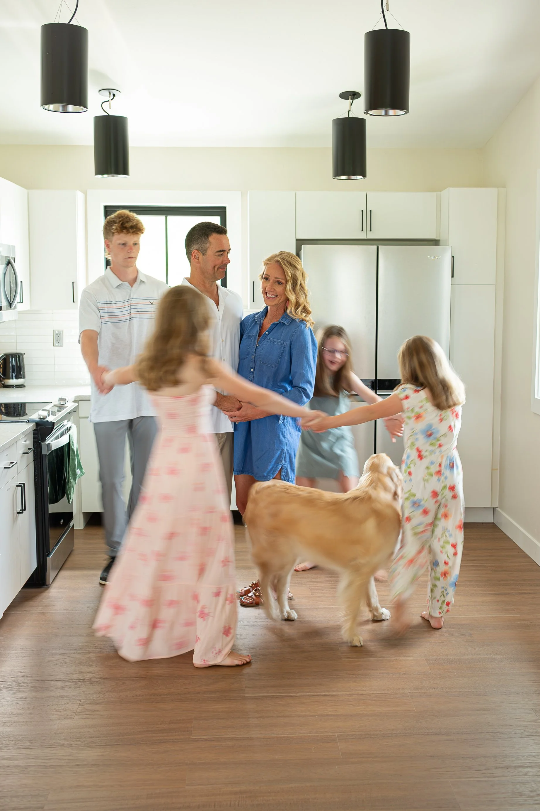 A family is gathered in their kitchen, smiling and holding hands in a circle around a large golden retriever dog. The scene appears lively and joyful, with children and adults participating in a group activity.