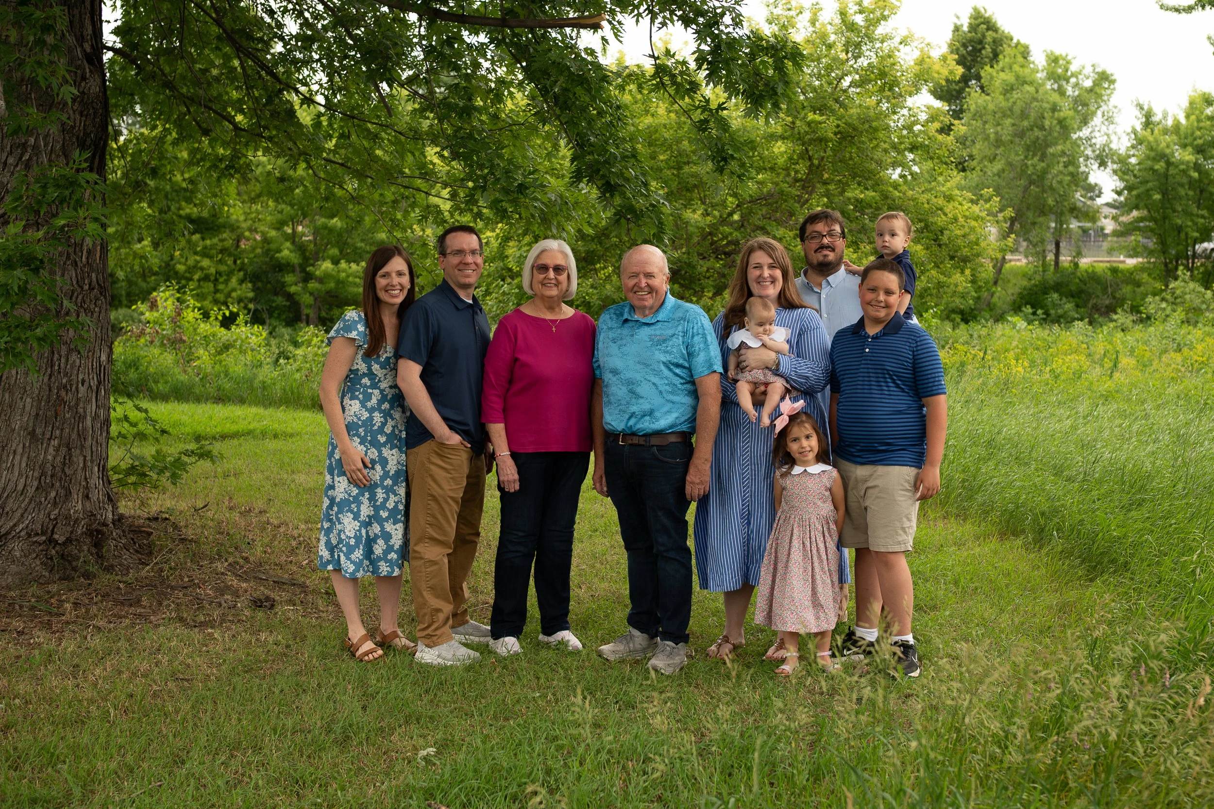 A multigenerational family of ten people standing outdoors under a large tree in a grassy area, smiling at the camera. There are three children, and the group includes elderly and middle-aged adults, all dressed casually.