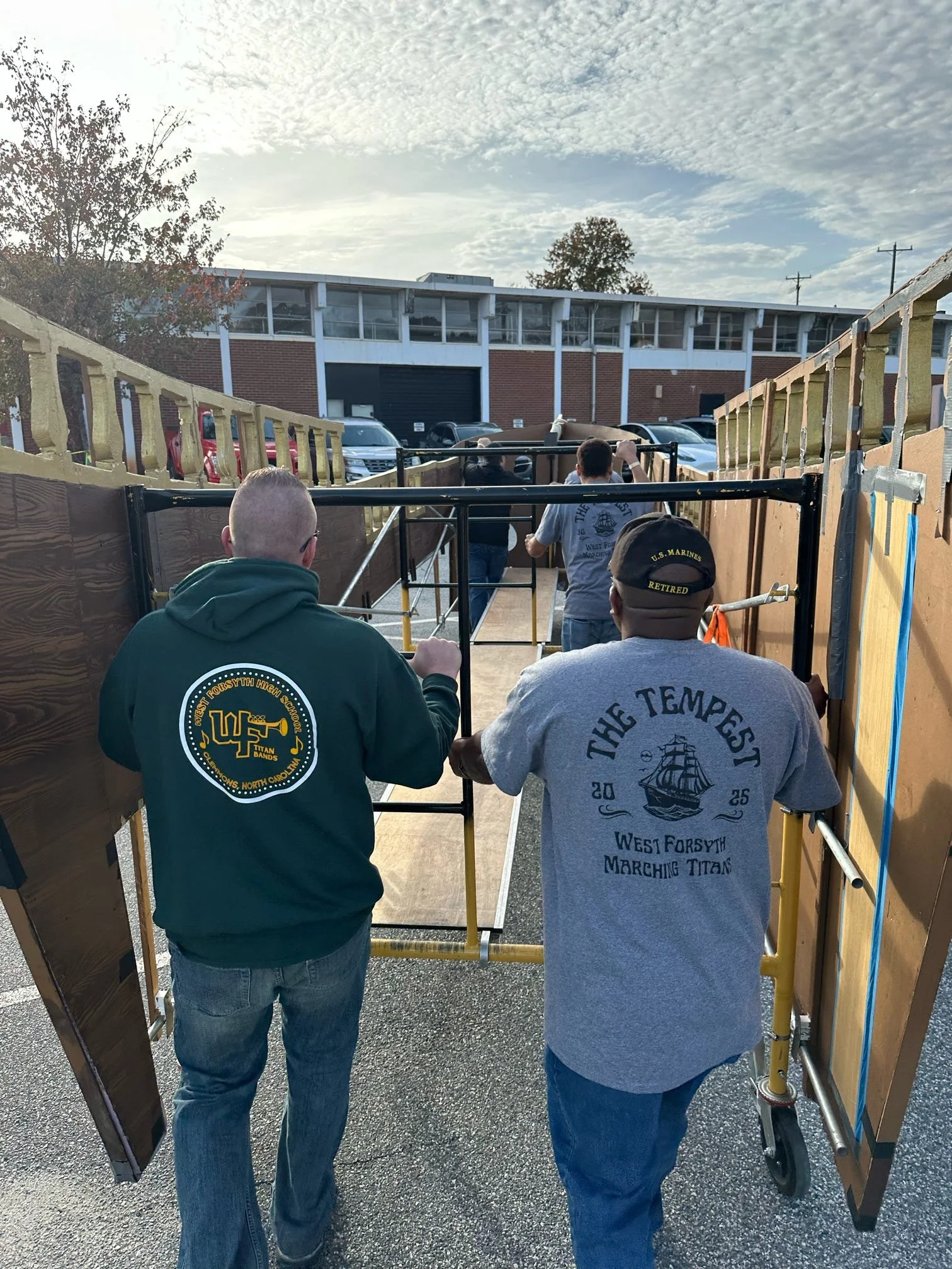 People walking through a passageway made of wooden panels and metal frames, with parked cars and a building in the background.