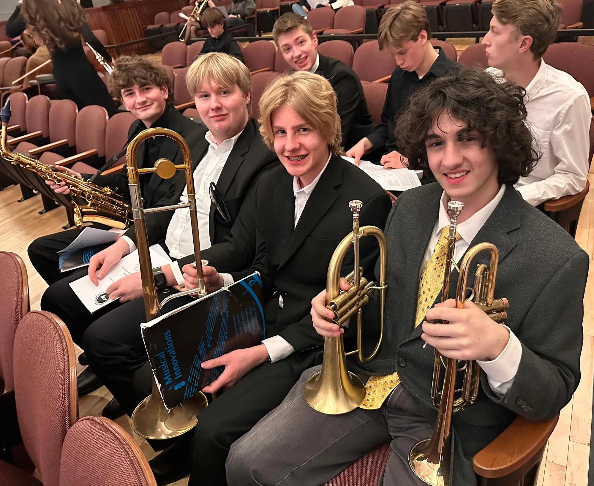A group of young musicians in formal attire sitting in auditorium seats, holding brass and wind instruments, smiling at the camera.