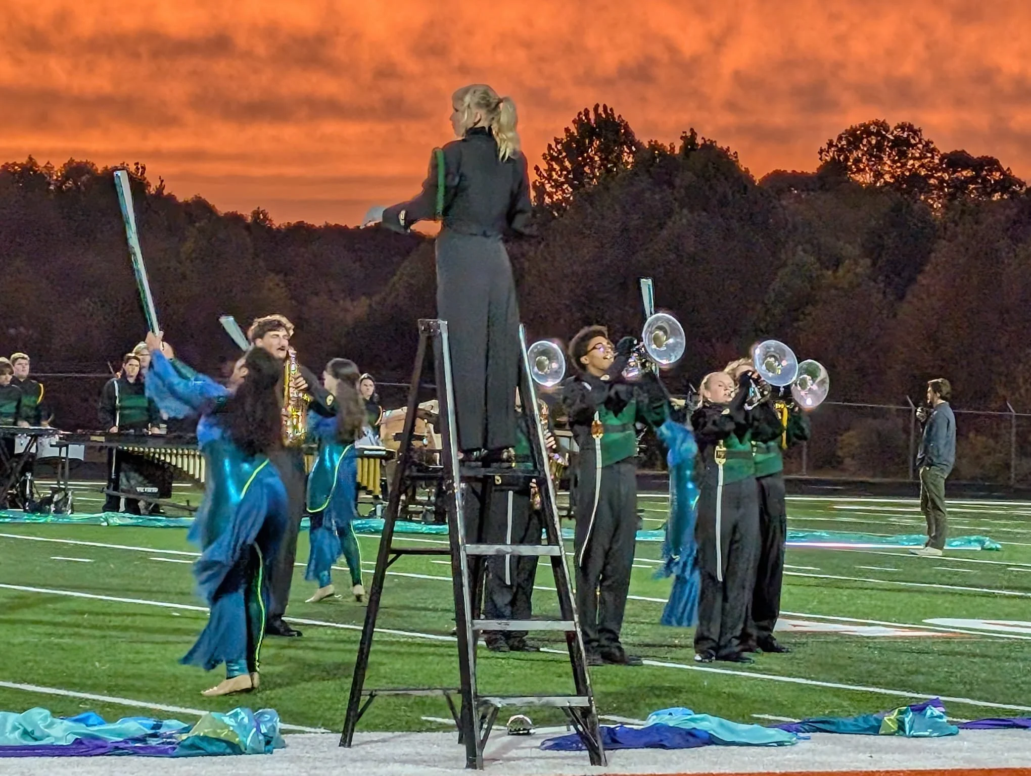 Band members in a marching band practice on a football field during sunset, with orange clouds in the sky and trees in the background.