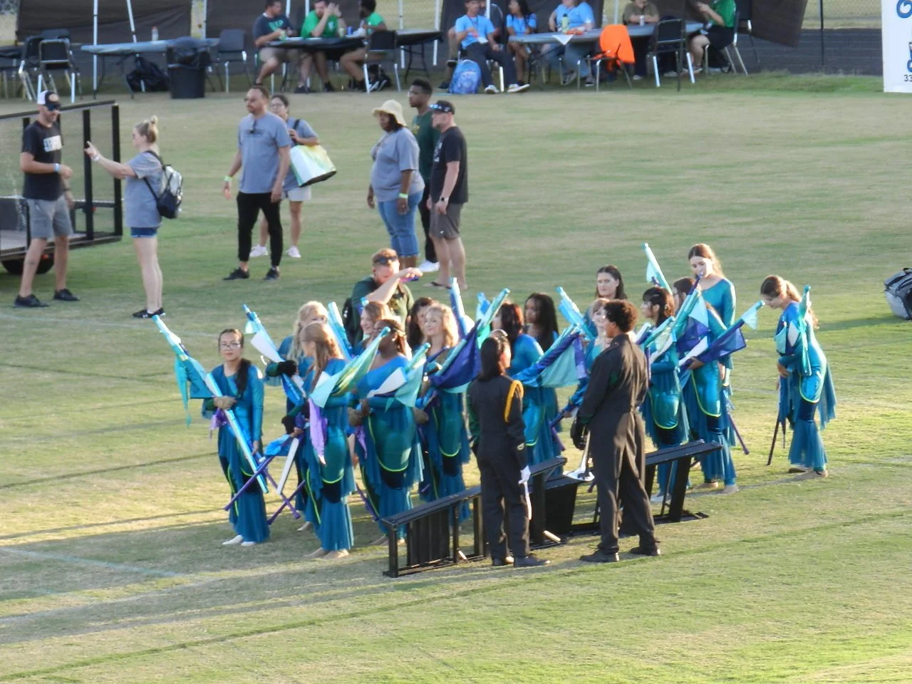 A group of women dressed in blue uniforms holding flags, gathered in a circle on a grassy field during a daytime outdoor event, with spectators and event organizers in the background.