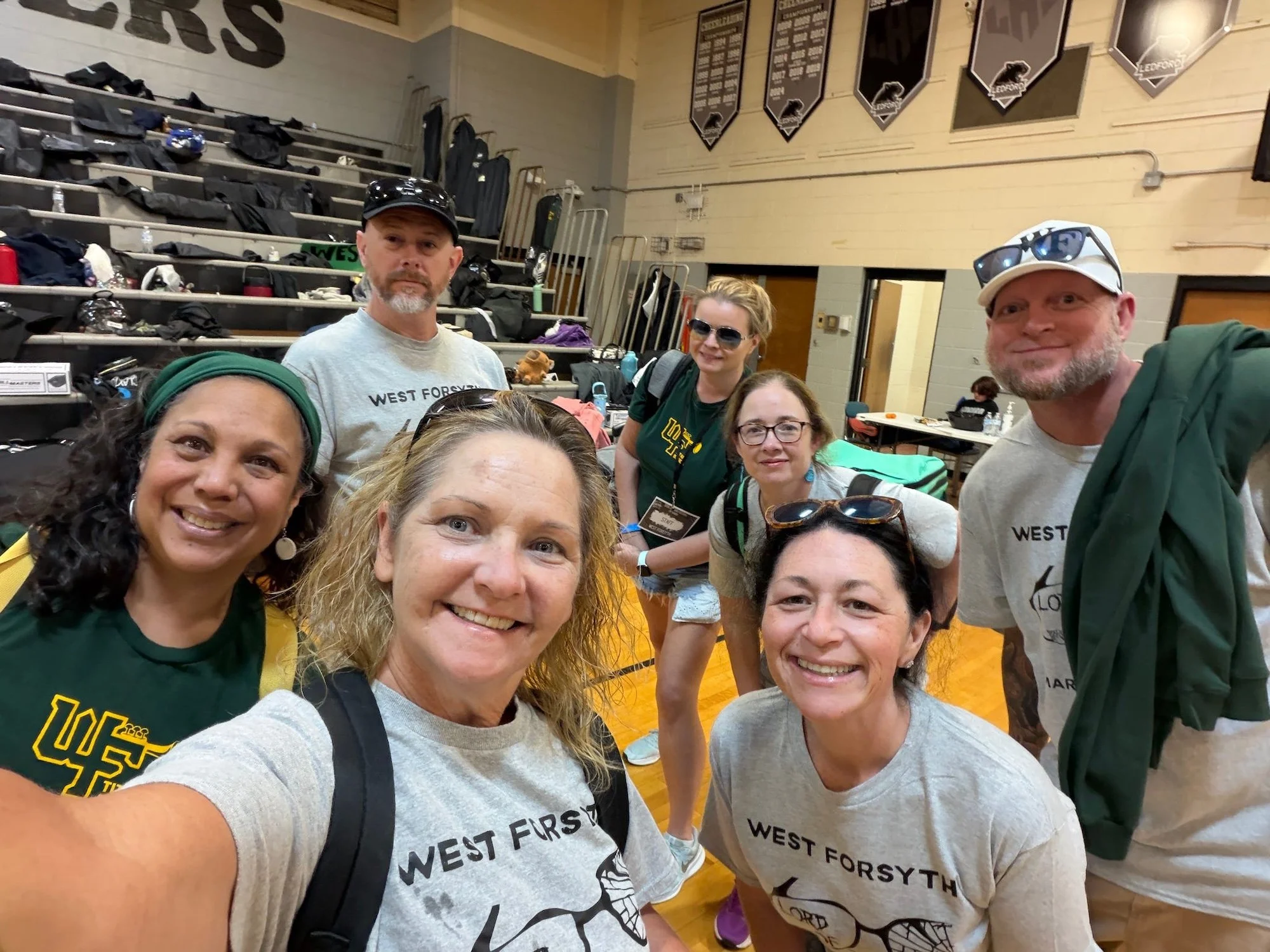 Group of five smiling adults taking a selfie in a gymnasium or sports hall, with bleachers and banners in the background, and sports equipment and bags on the bleachers.