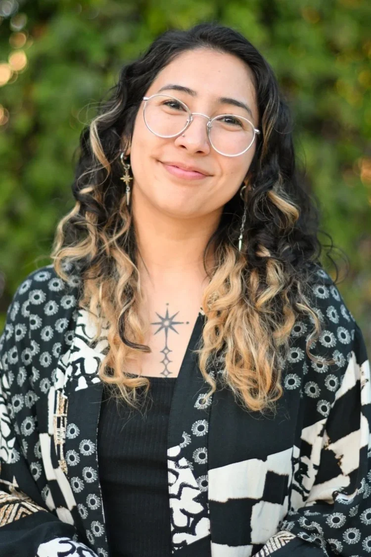Headshot of a woman with glasses, dark curly hair, and chest tattoos smiling gently