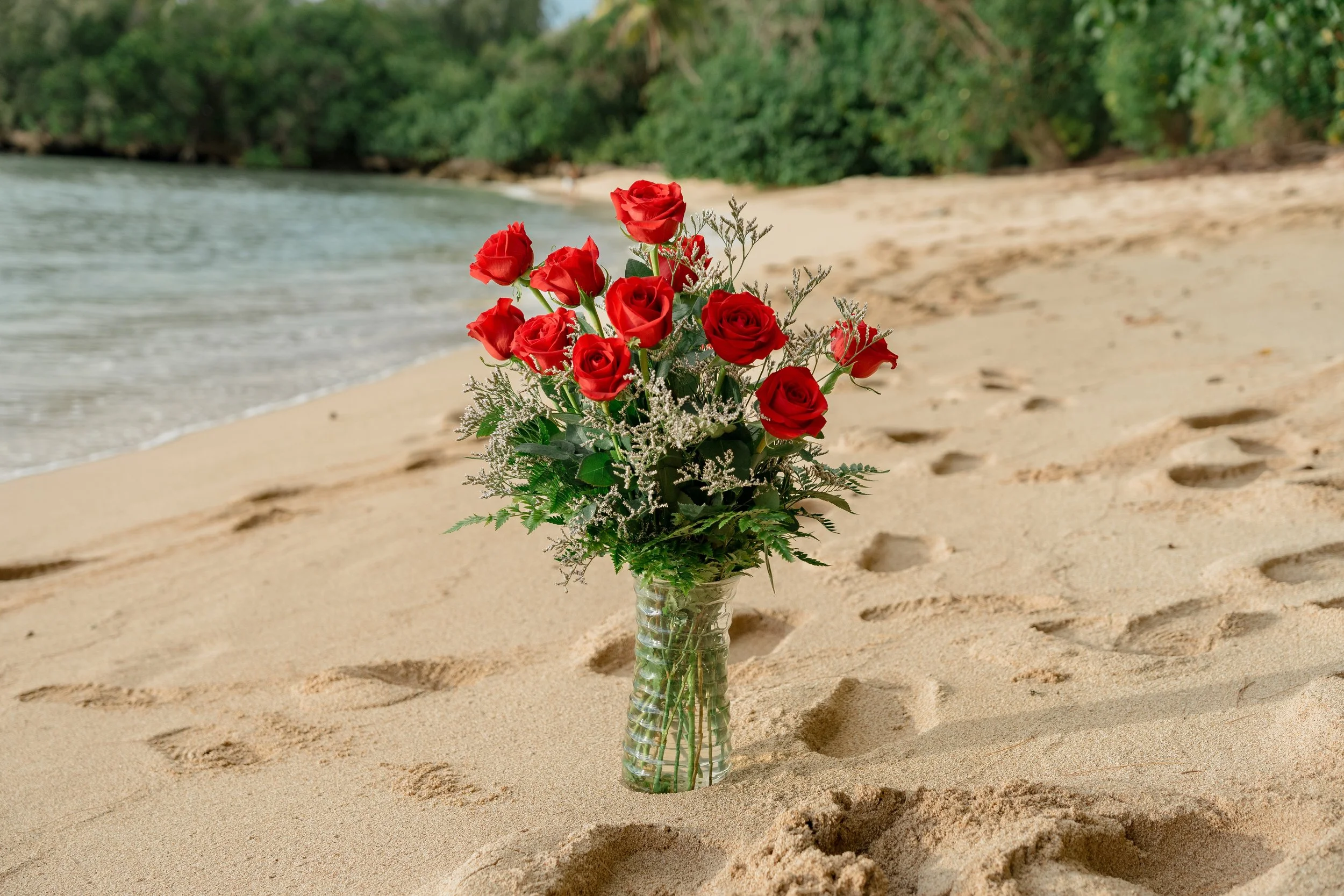 A bouquet of red roses in a clear glass vase placed on sandy beach with footprints, near water and green trees in the background.