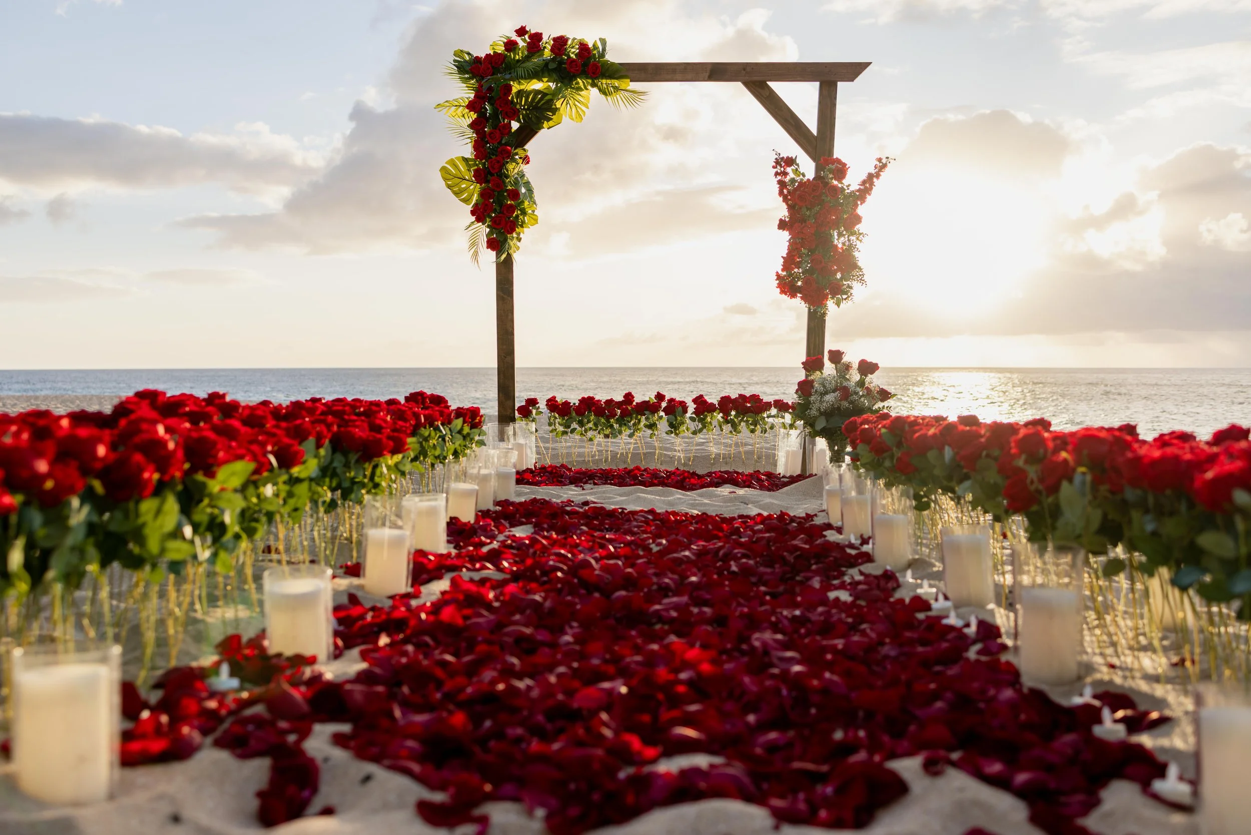 Hawaii beach proposal setup at sunset, featuring a rose-petal aisle, candle-lined pathway, and a custom floral arch overlooking the ocean.