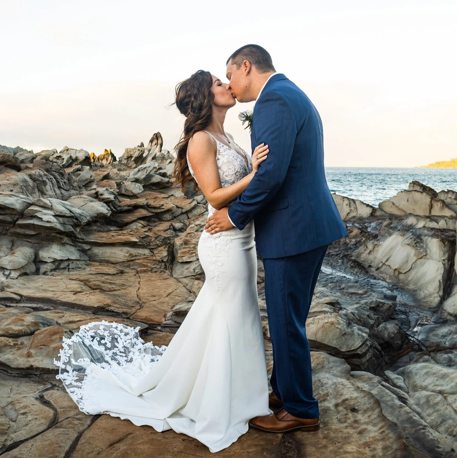 A bride and groom kiss on a rocky shoreline during a wedding photo shoot.