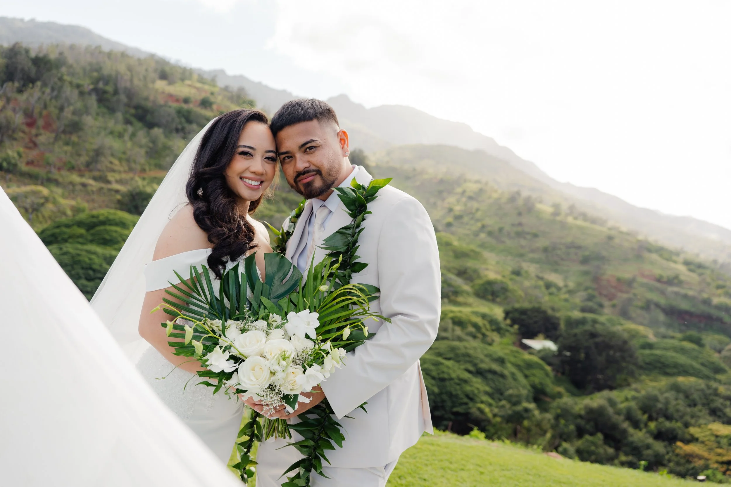 A newlywed couple in wedding attire standing outdoors with green mountains in the background. The bride is holding a bouquet of white flowers and wearing an off-shoulder white wedding dress. The groom is in a white suit with a green leafy garland around his neck. They are smiling and close together.
