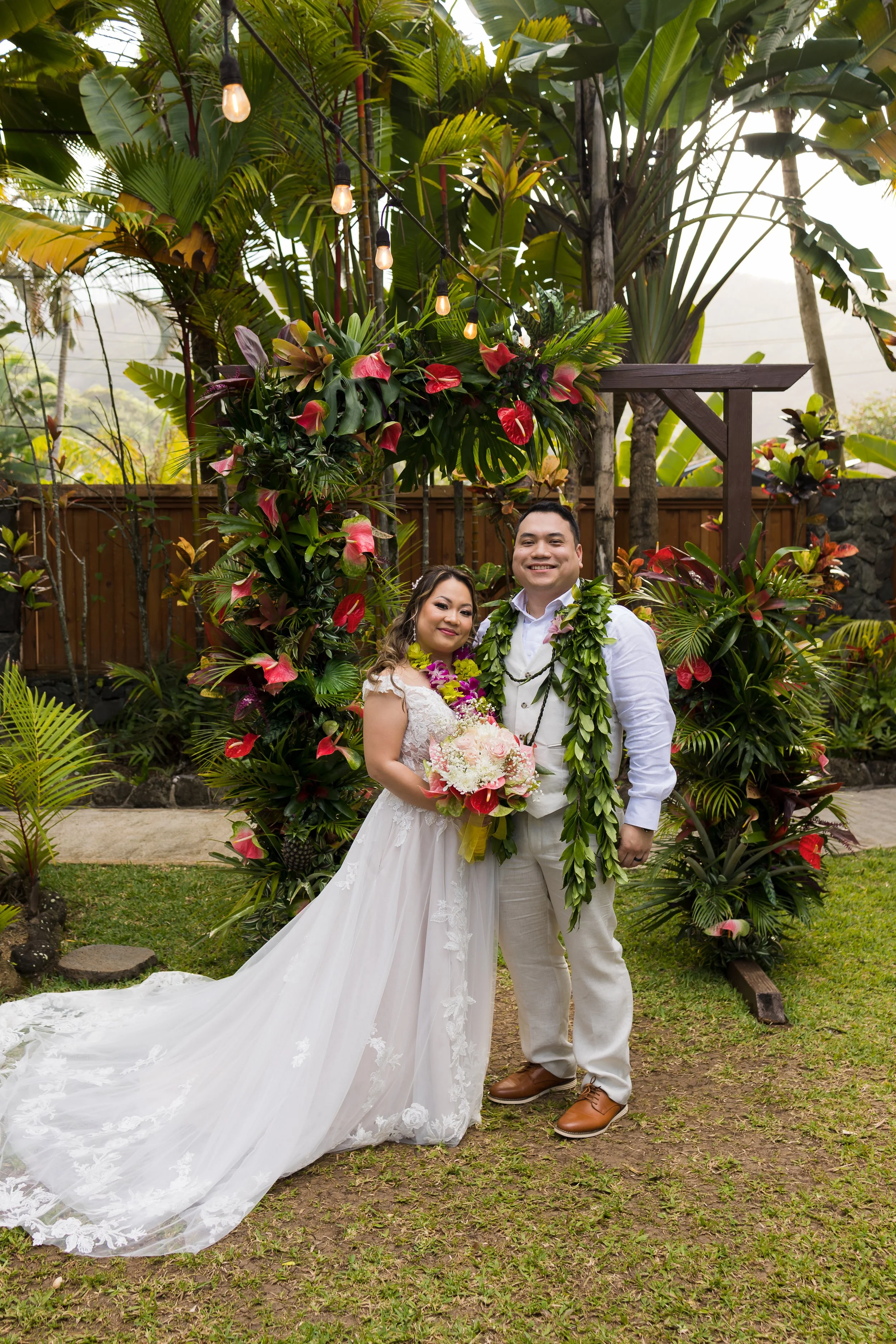 A newlywed couple stands outdoors in front of a floral arch, with lush tropical plants and string lights overhead. The bride wears a white wedding gown, holds a bouquet of flowers, and has floral leis around her neck. The groom wears a white shirt, light-colored pants, a lei, and smiles at the camera.