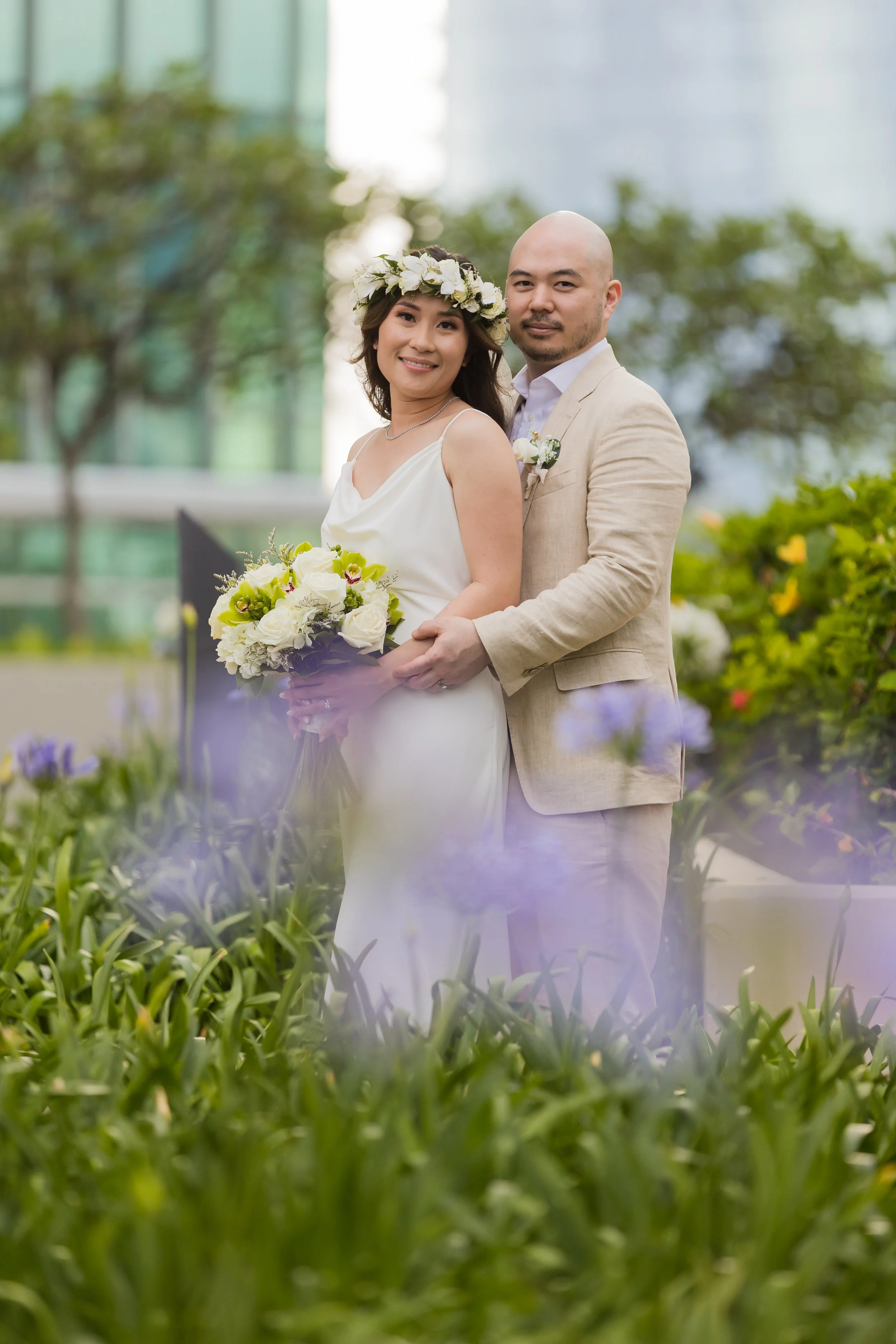 A bride and groom standing outdoors in a garden, smiling at the camera. The bride wears a white dress with a floral crown and holds a bouquet of white and green flowers. The groom wears a beige suit and white shirt.