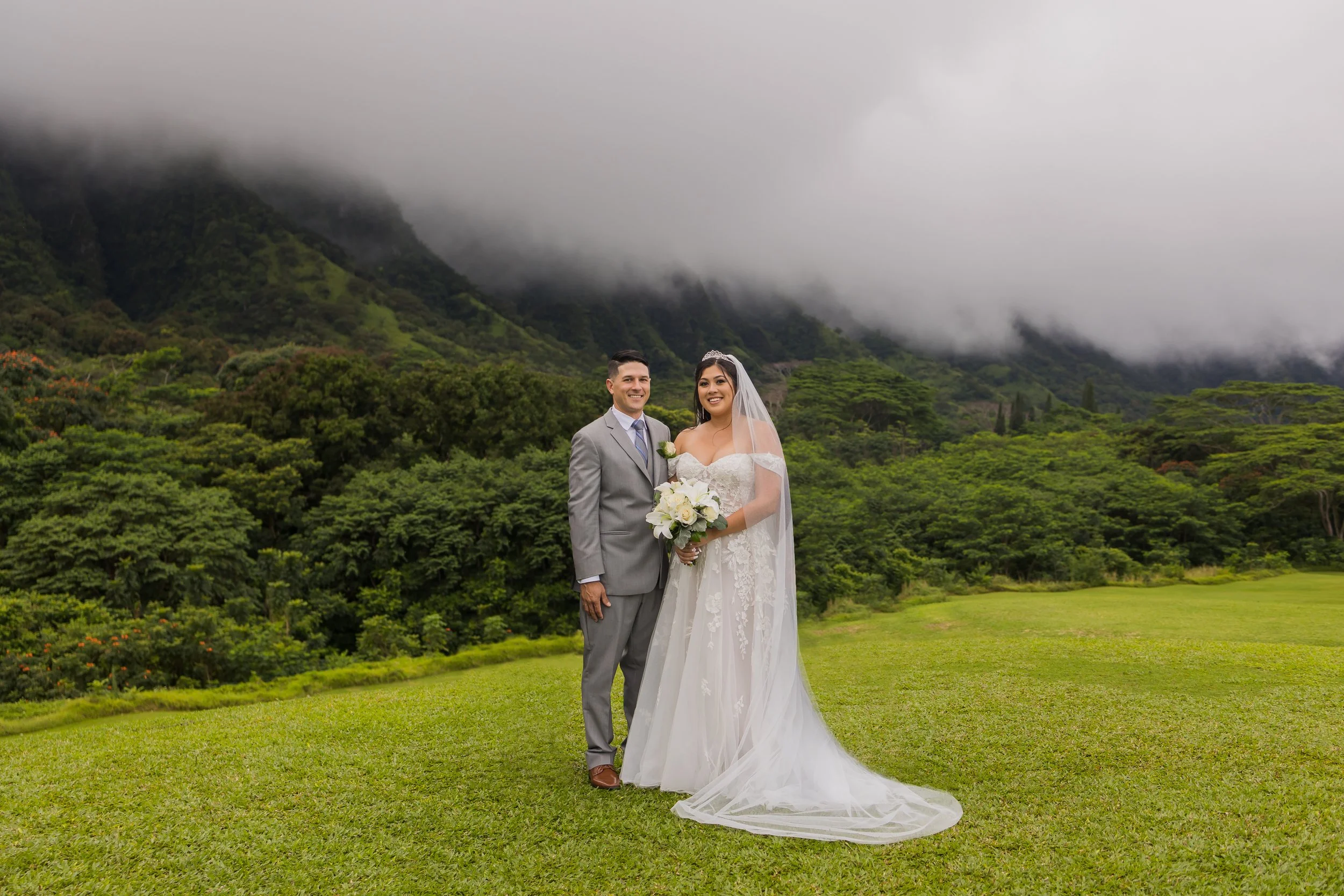 A bride and groom standing together on a lush green lawn with mountains and low-hanging clouds in the background.