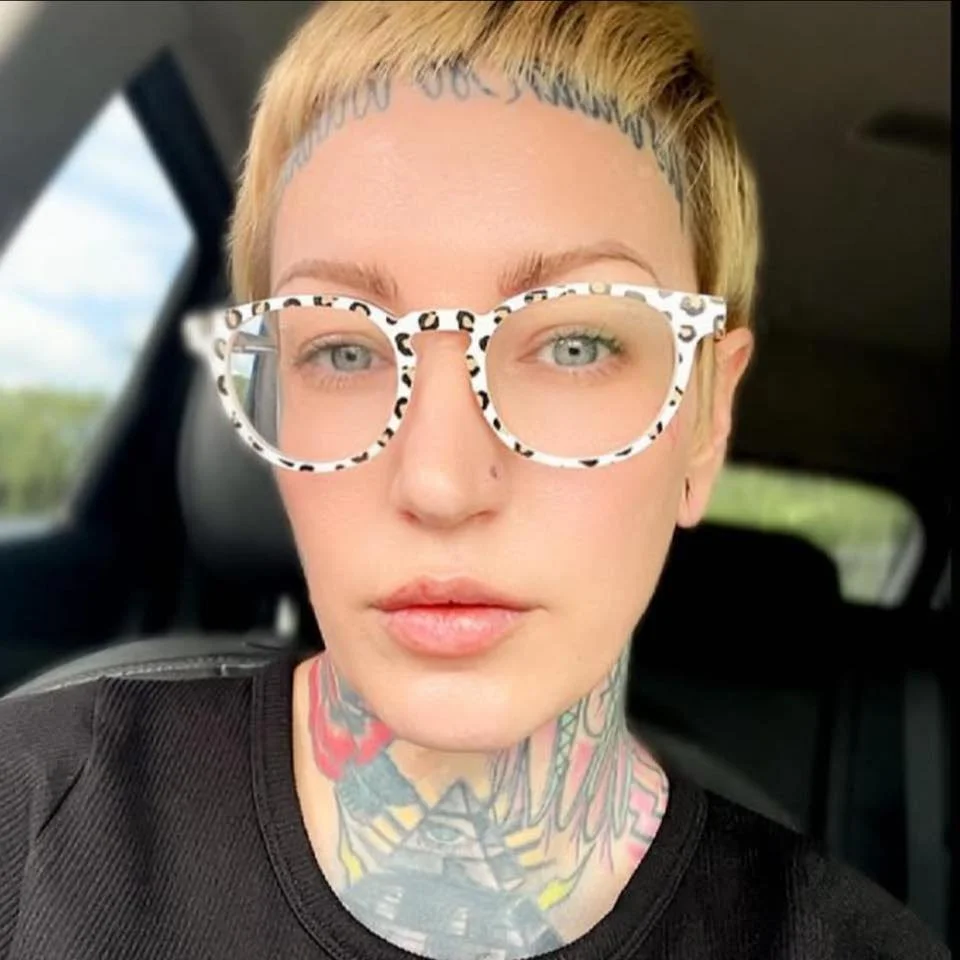 A person with short blond hair wearing white-framed glasses with a leopard print pattern, tattoos on their neck, and a black shirt, sitting inside a car.
