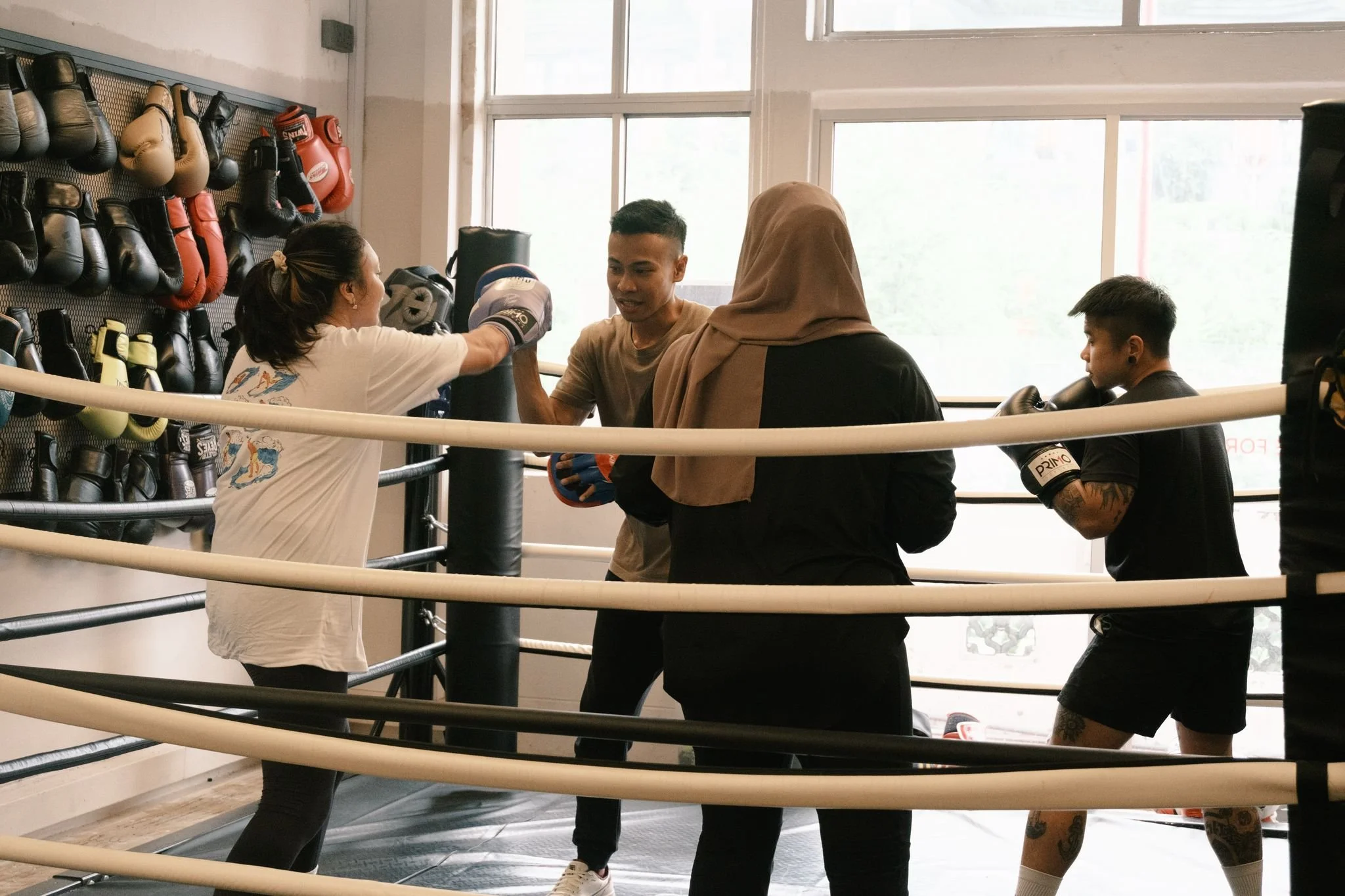 A boxing training session in a gym with four people, including a female trainer sparring with a male boxer, while two other women observe. The trainer and boxer are wearing boxing gloves, and boxing equipment is visible on the wall.