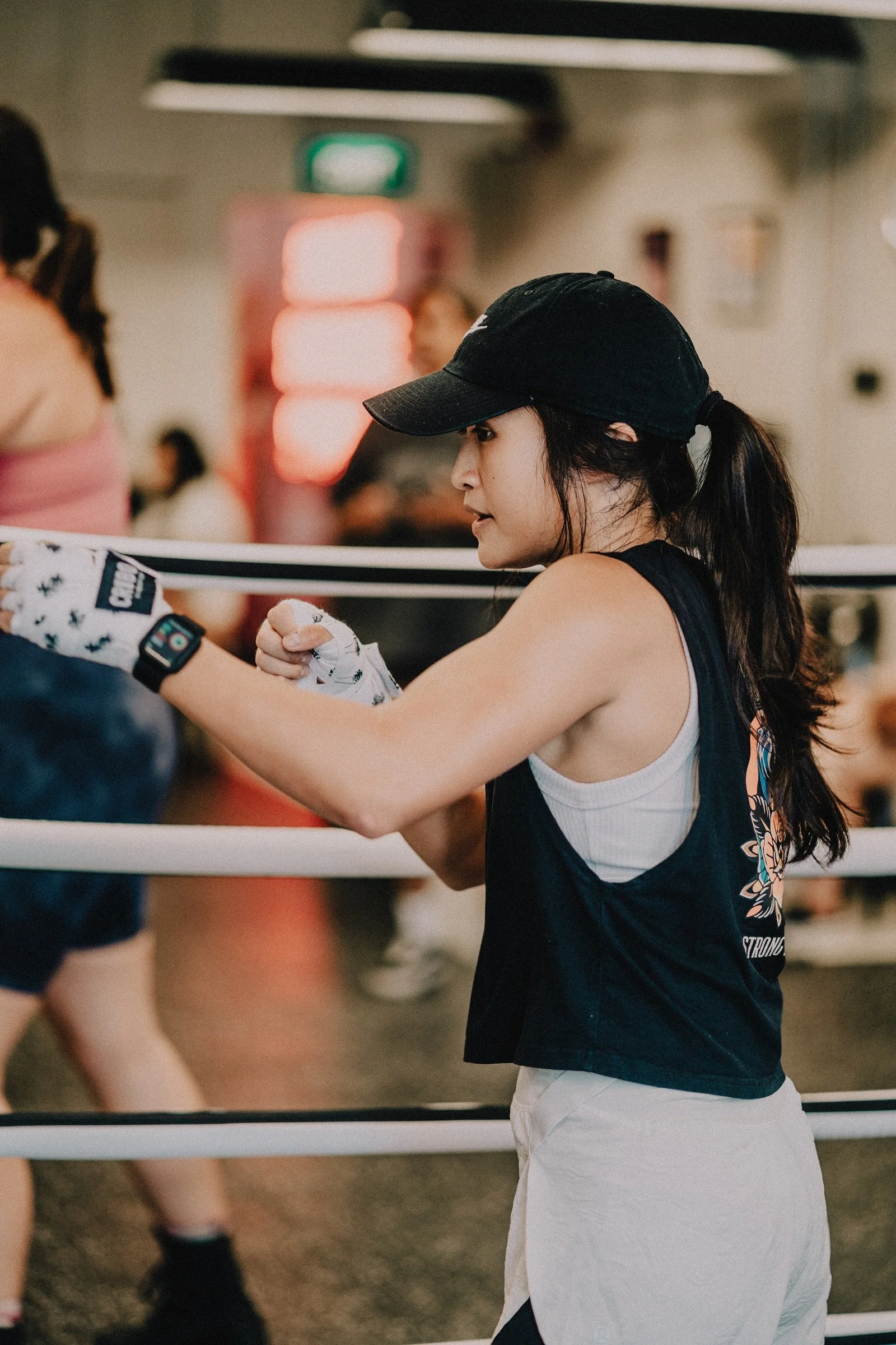 Woman in a black cap and white hand wraps holding her guard beside the boxing ring during a group class at Sweat Science Studio, Clarke Quay.