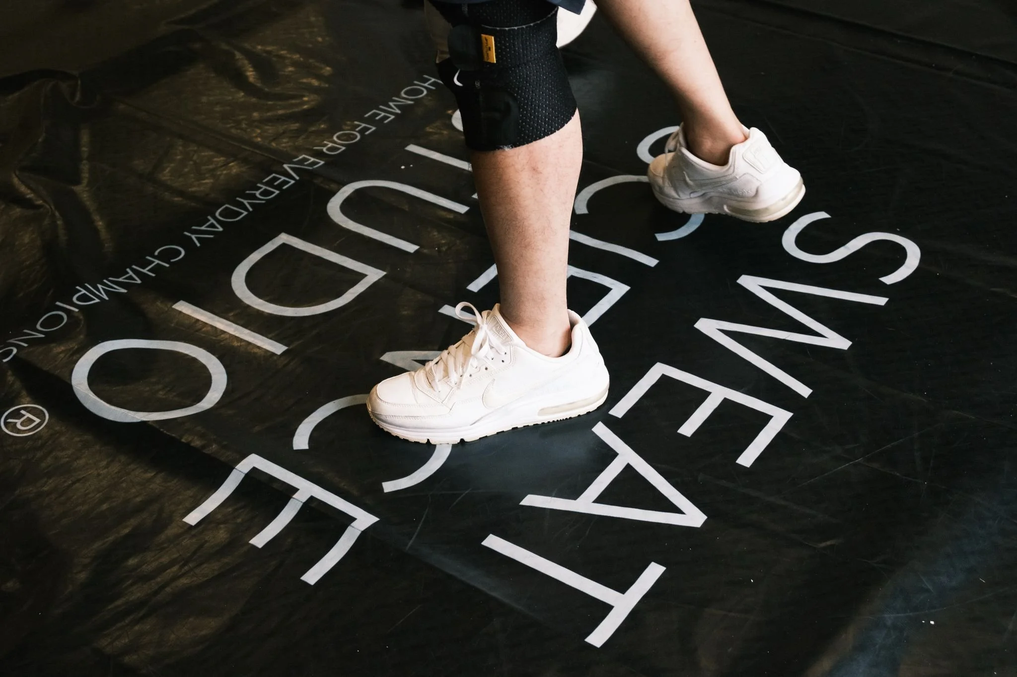 A person wearing white sneakers and black shorts standing on a black floor with white printed text, including the words 'FALL' and 'S' and other partially visible letters.