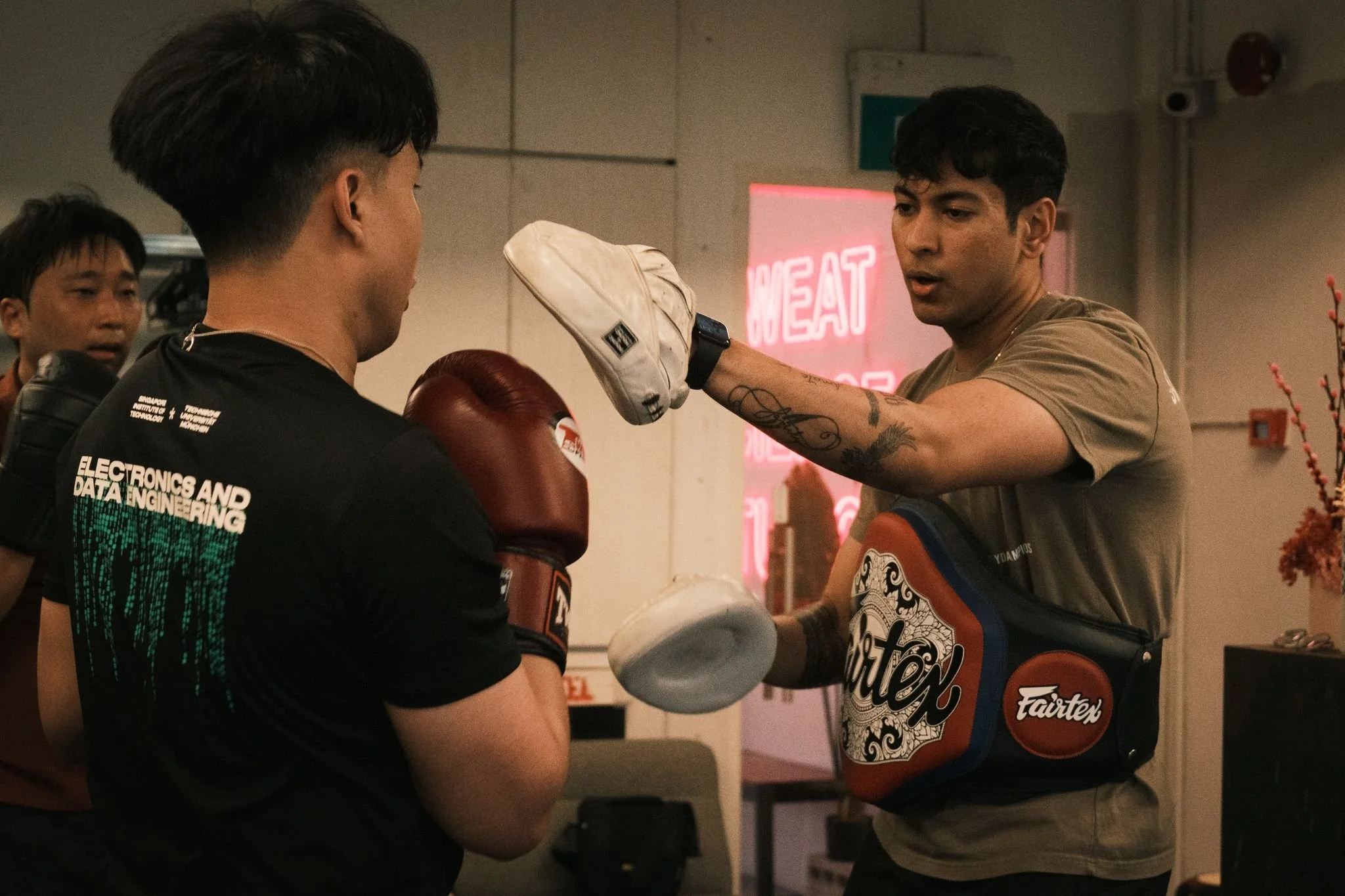 An indoor scene with two men training in boxing. One man is holding a padded focus mitt while the other is wearing boxing gloves and is sparring. There are other people observing in the background, and a neon sign that reads "MEAT" is visible on the wall.