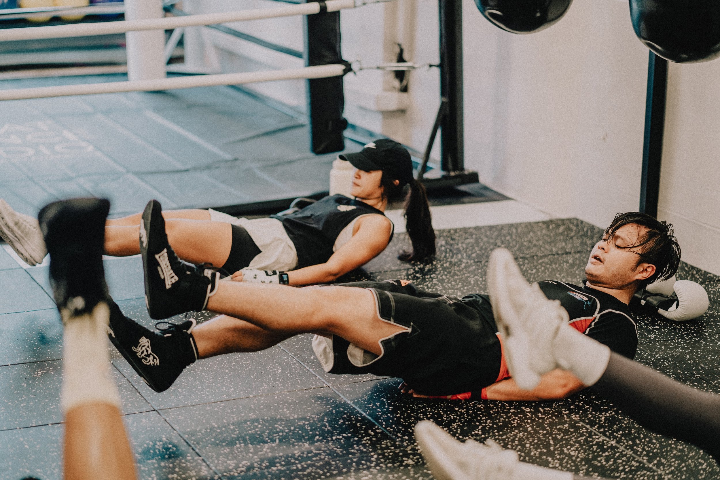 Two members doing flutter kicks on the gym floor in boxing gear with a boxing ring in the background at Sweat Science Studio Singapore