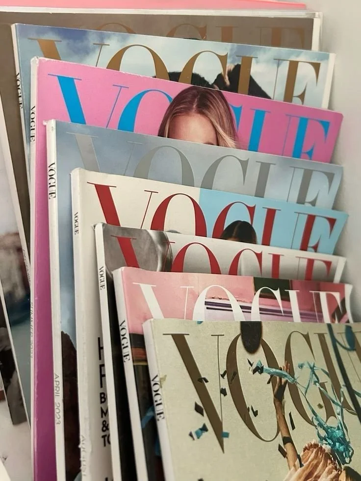 A collection of Vogue magazines stacked on top of each other with colorful covers, including pink, blue, and beige, featuring various models and fashion-related images.