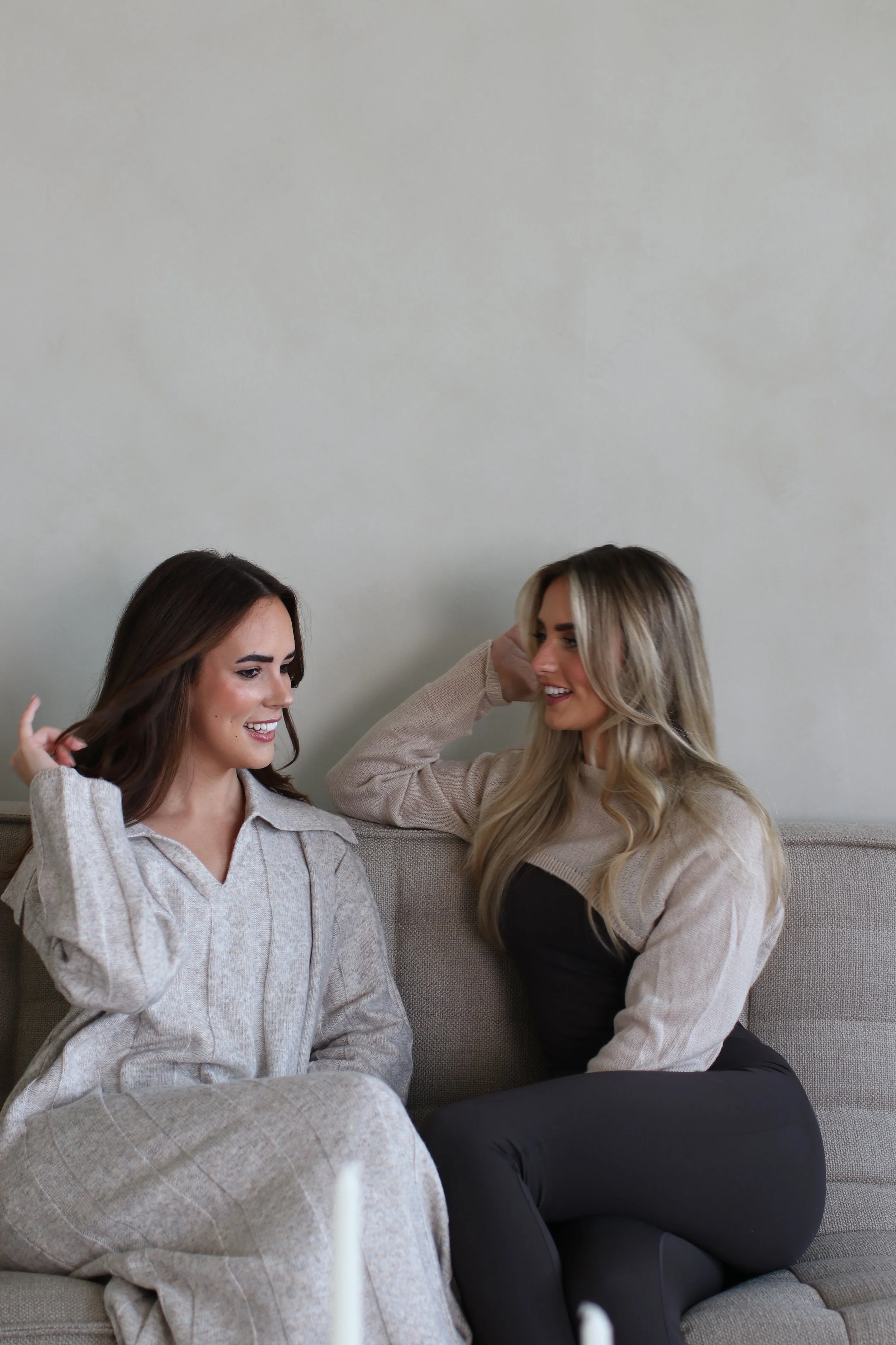 Two women sitting on a beige couch, smiling and talking while one touches her hair. Both dressed in neutral-colored clothing, with a plain light wall in the background.